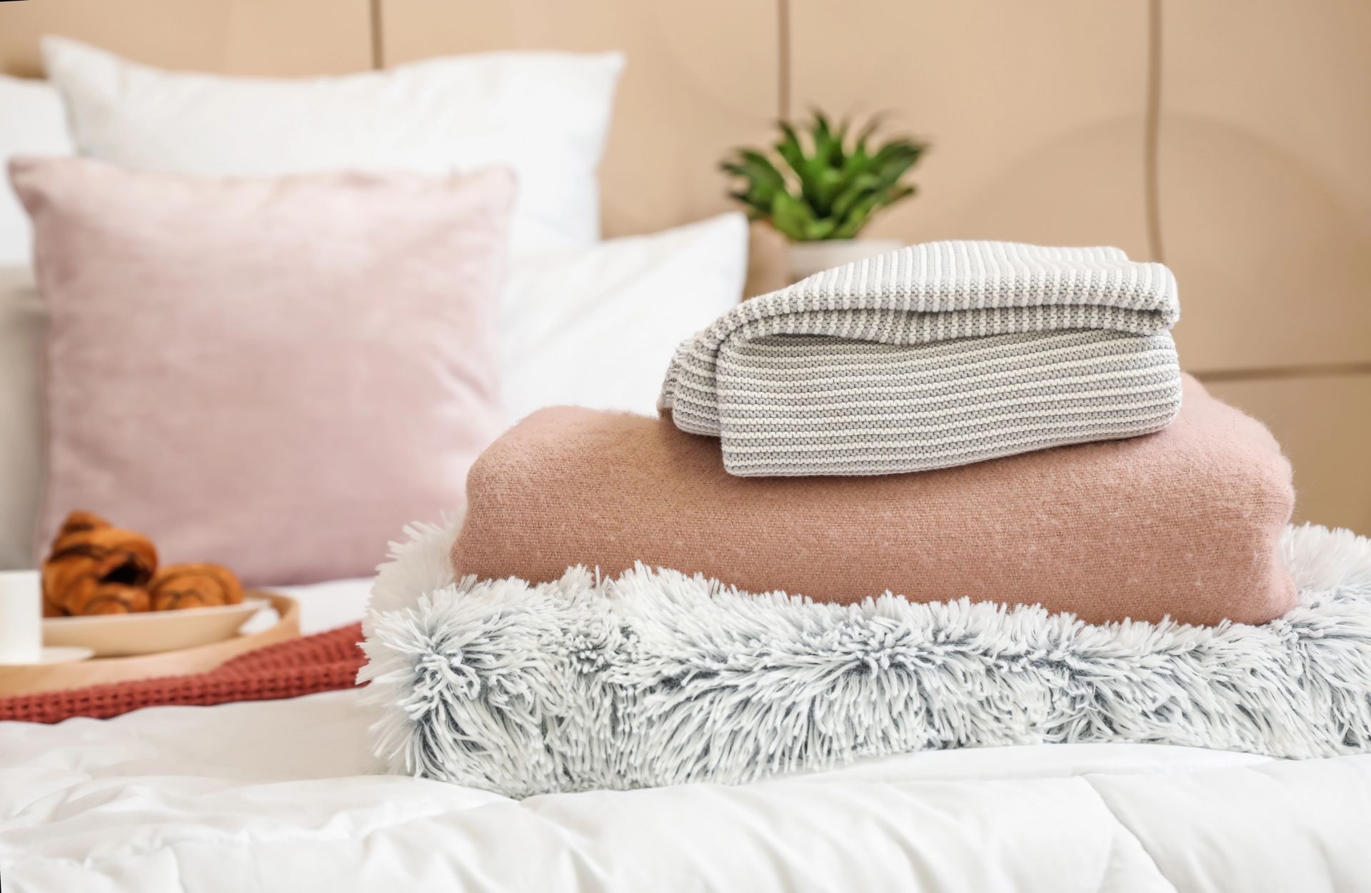 Coperte di maglia piegate su un letto con cuscini rosa e una piccola pianta sullo sfondo.