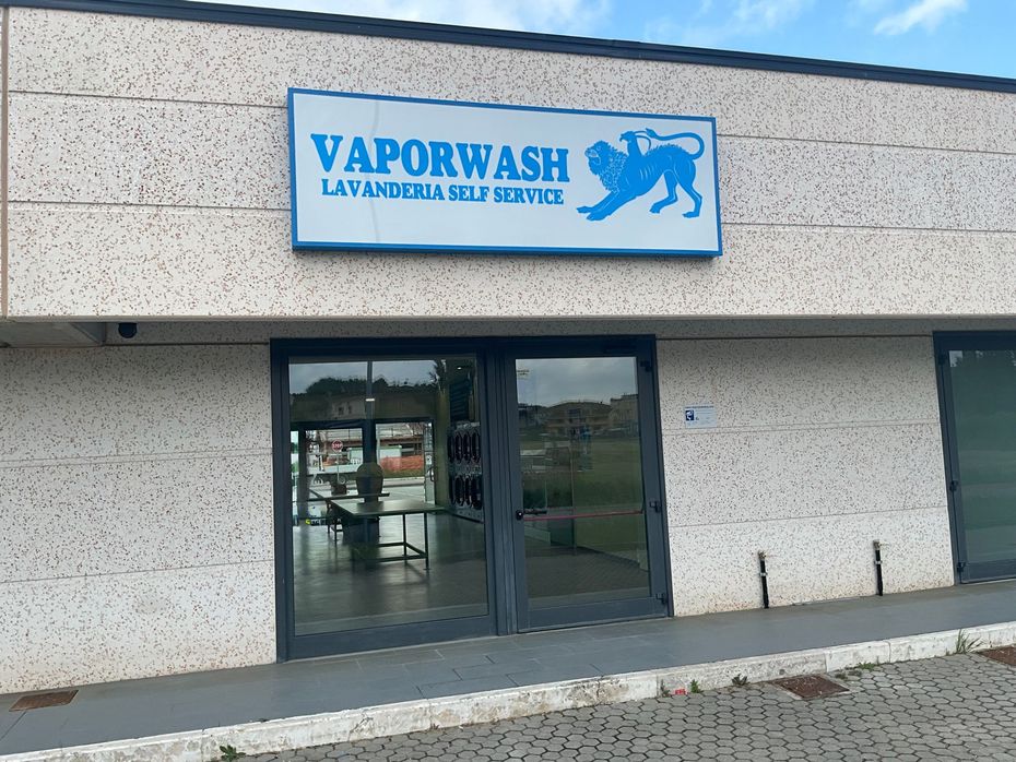 Lavanderia a gettoni Vaporwash con due porte a vetri, insegna blu e ingresso aperto su una strada soleggiata.