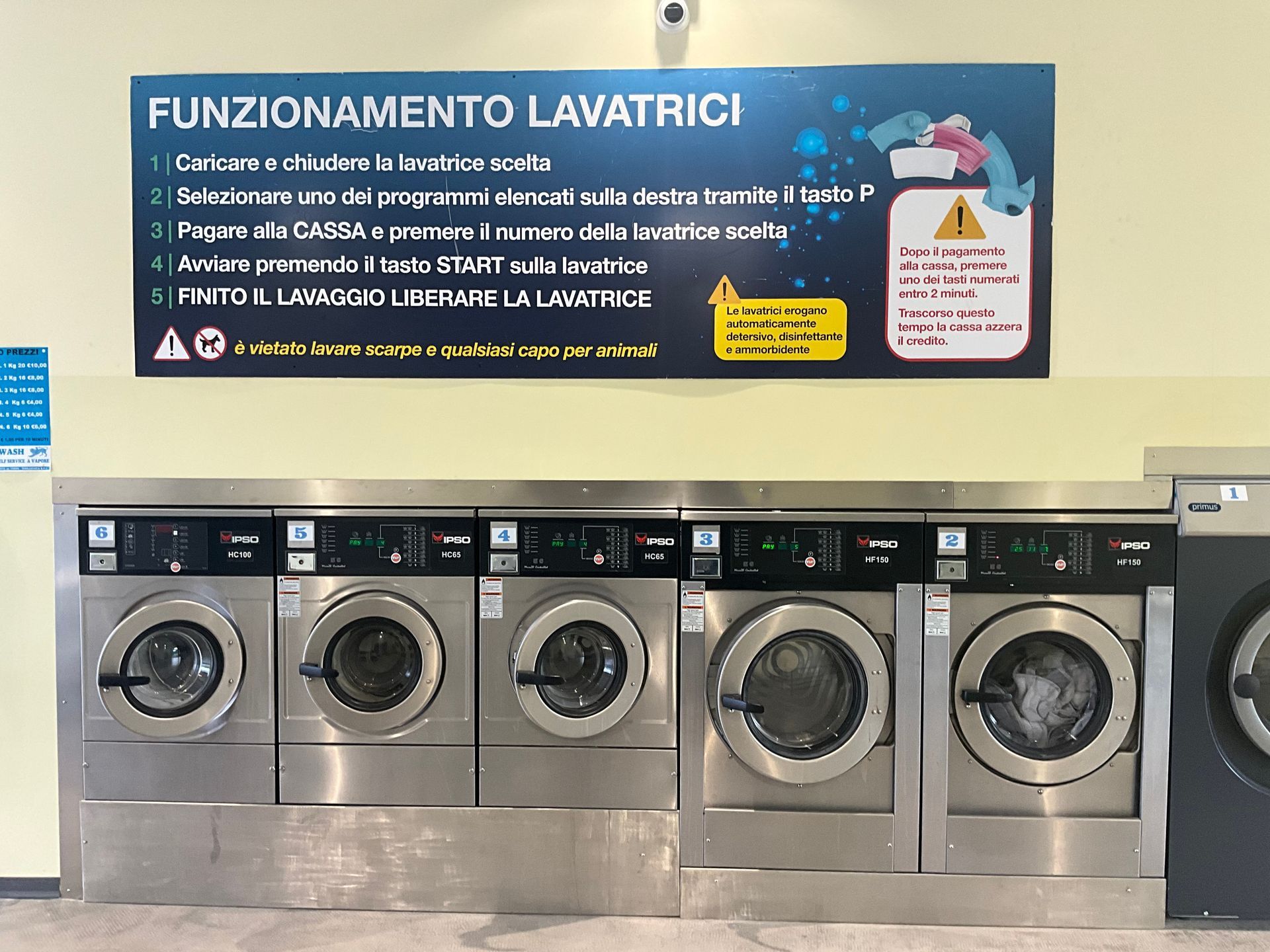 Una fila di lavatrici a carica frontale sotto un cartello con le istruzioni in italiano in una lavanderia a gettoni.