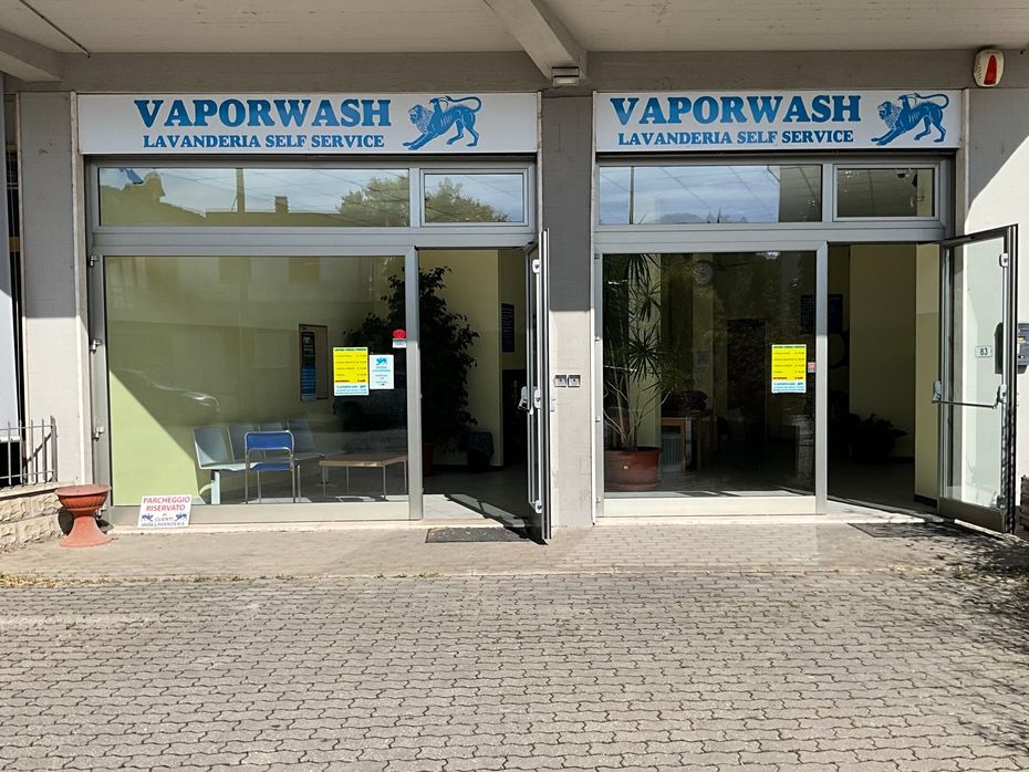 Lavanderia a gettoni Vaporwash con due porte a vetri, insegna blu e ingresso aperto su una strada soleggiata.