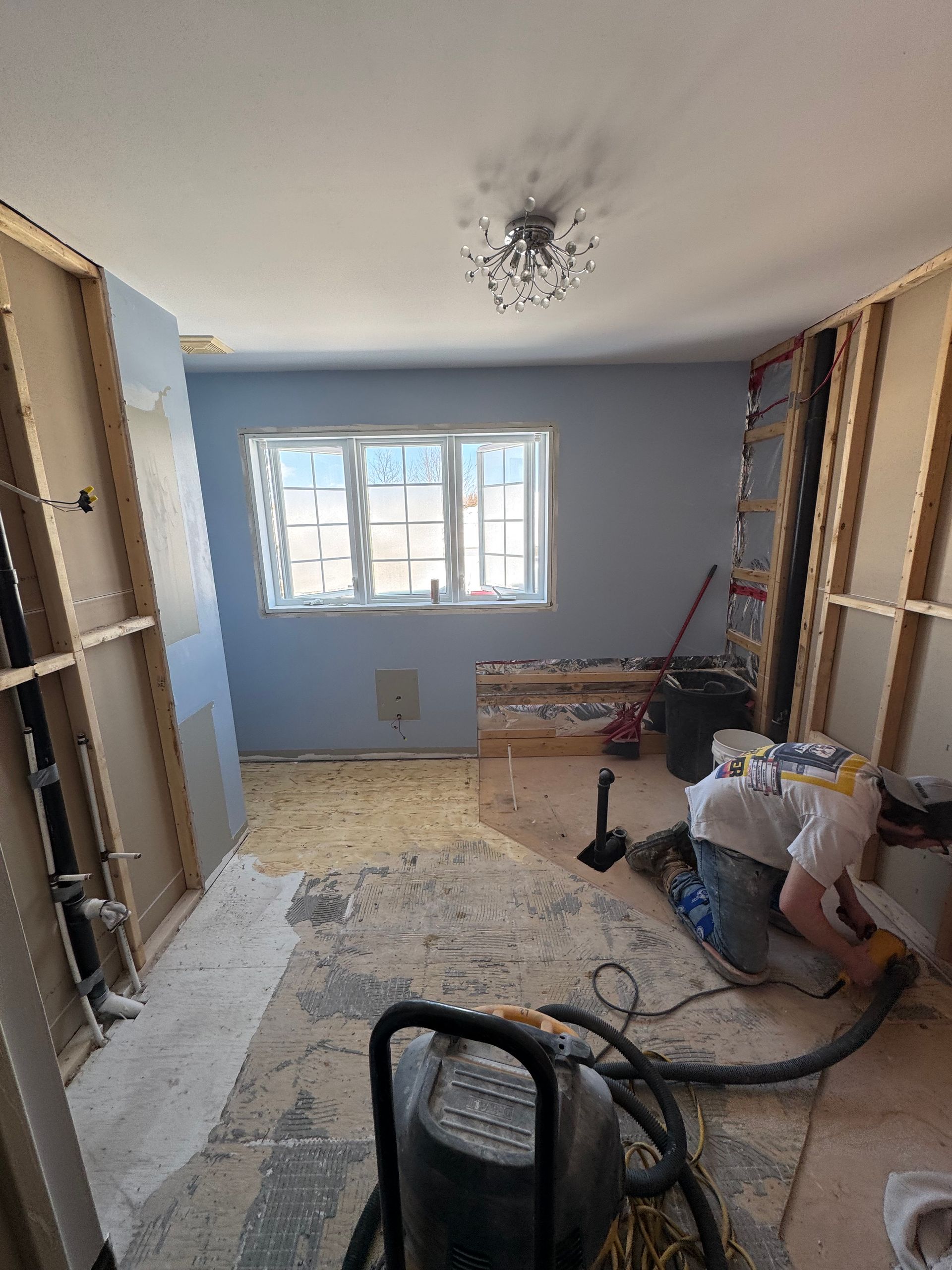 Pièce en rénovation ; murs apparents, murs bleus, fenêtre, ouvrier, aspirateur, débris au sol.