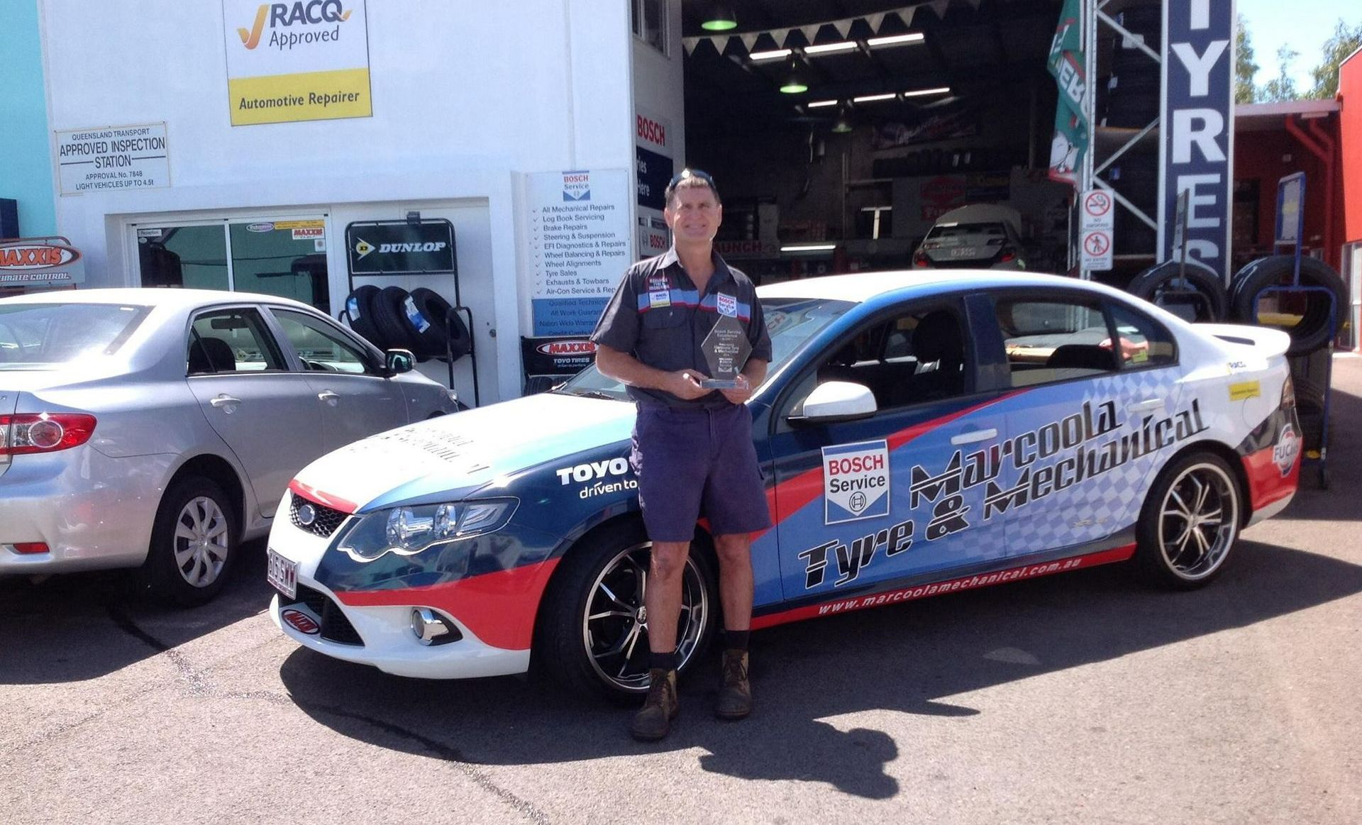 A Man Is Standing In Front Of A Car With A Trophy In His Hand — Marcoola Tyre & Mechanical In Marcoola, QLD
