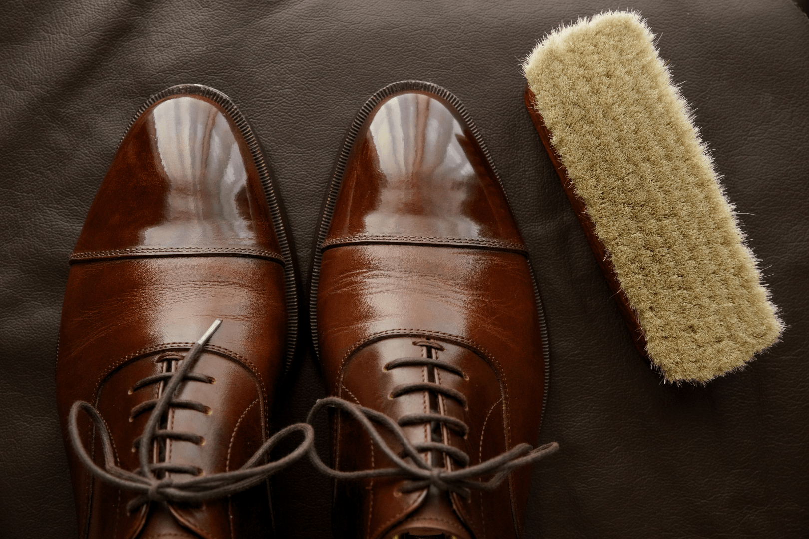 A pair of brown shoes next to a brush on a brown surface.