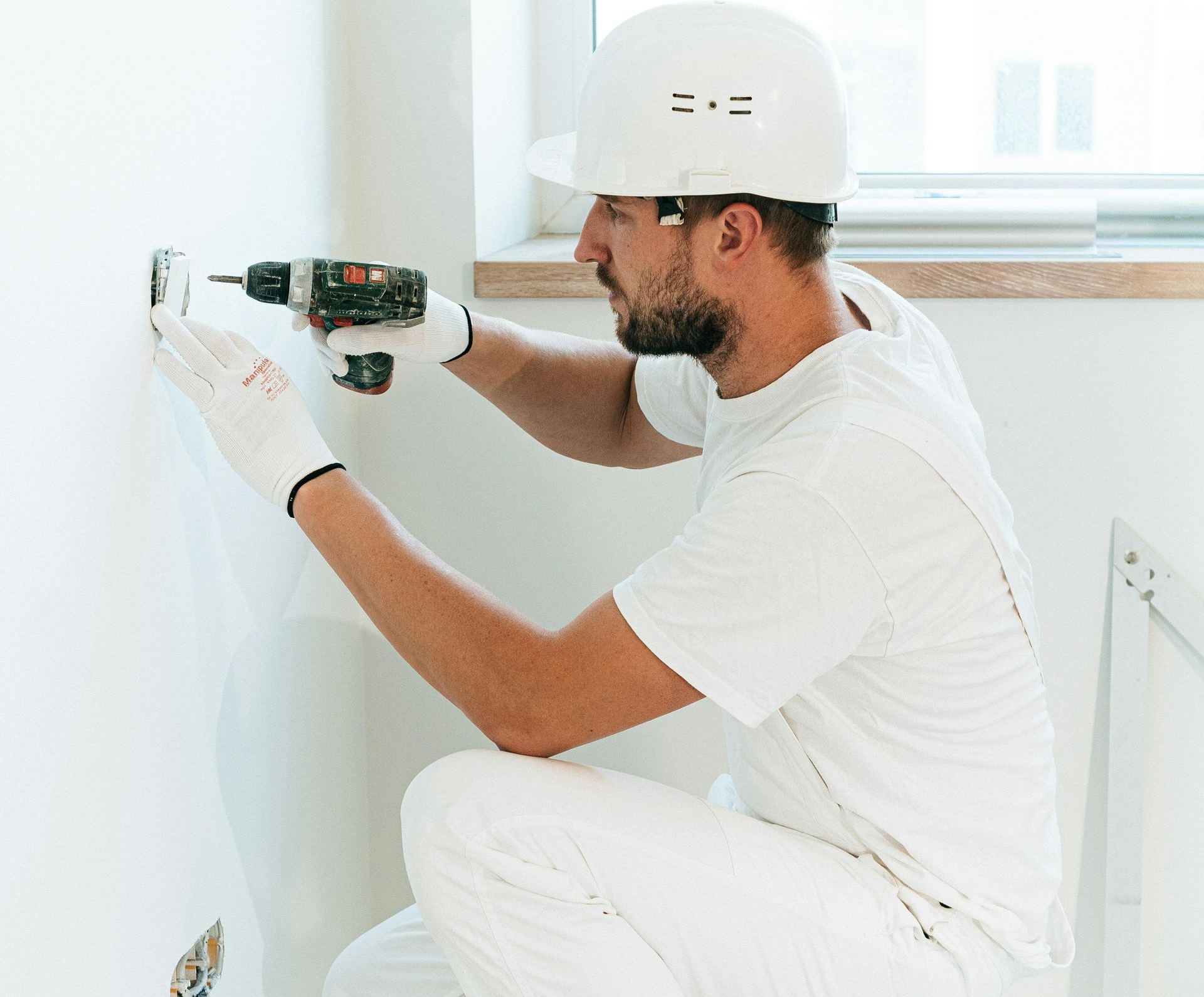 Worker in white clothing drilling into a wall in a bright room