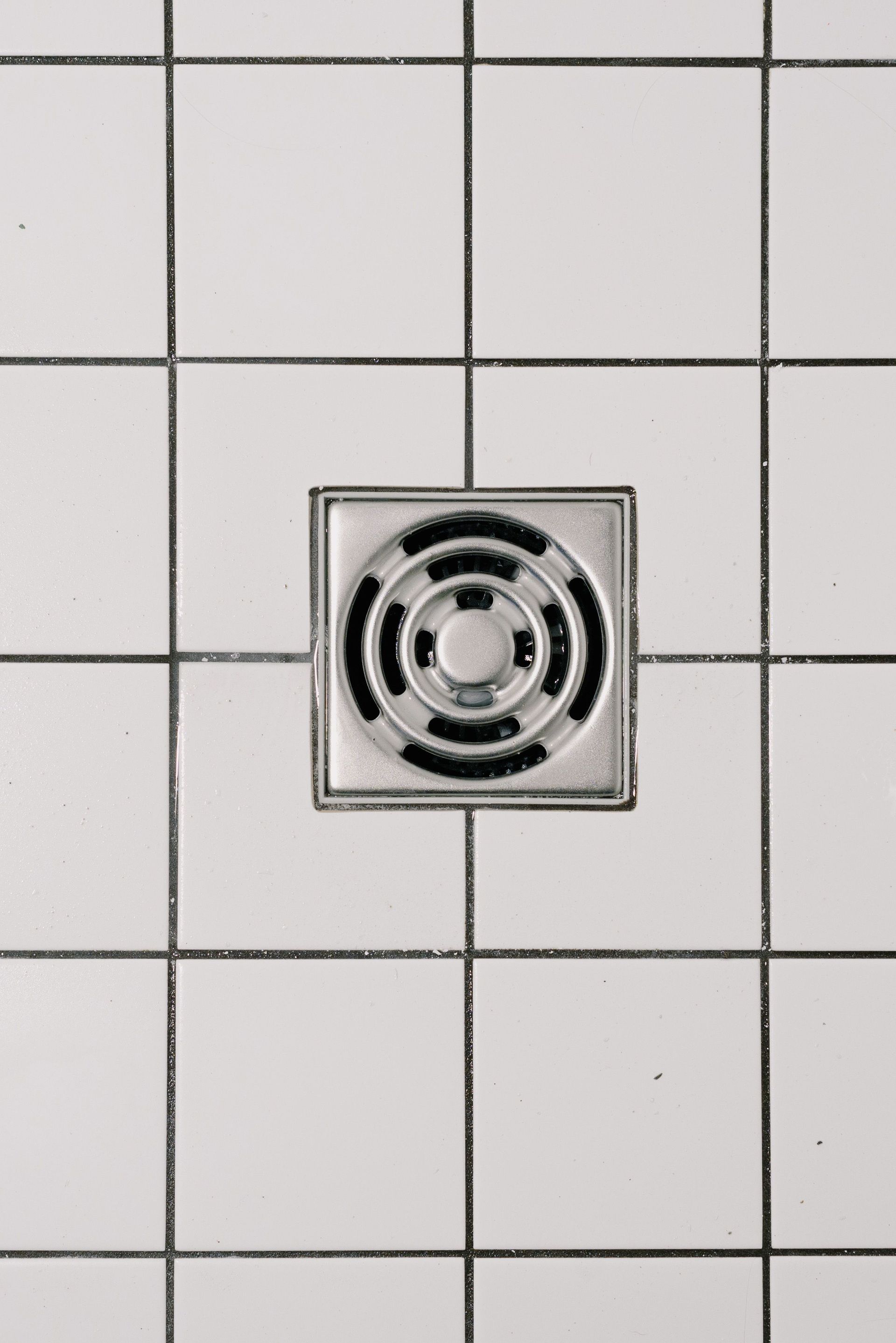 Square metal shower drain centered in white tiled floor.
