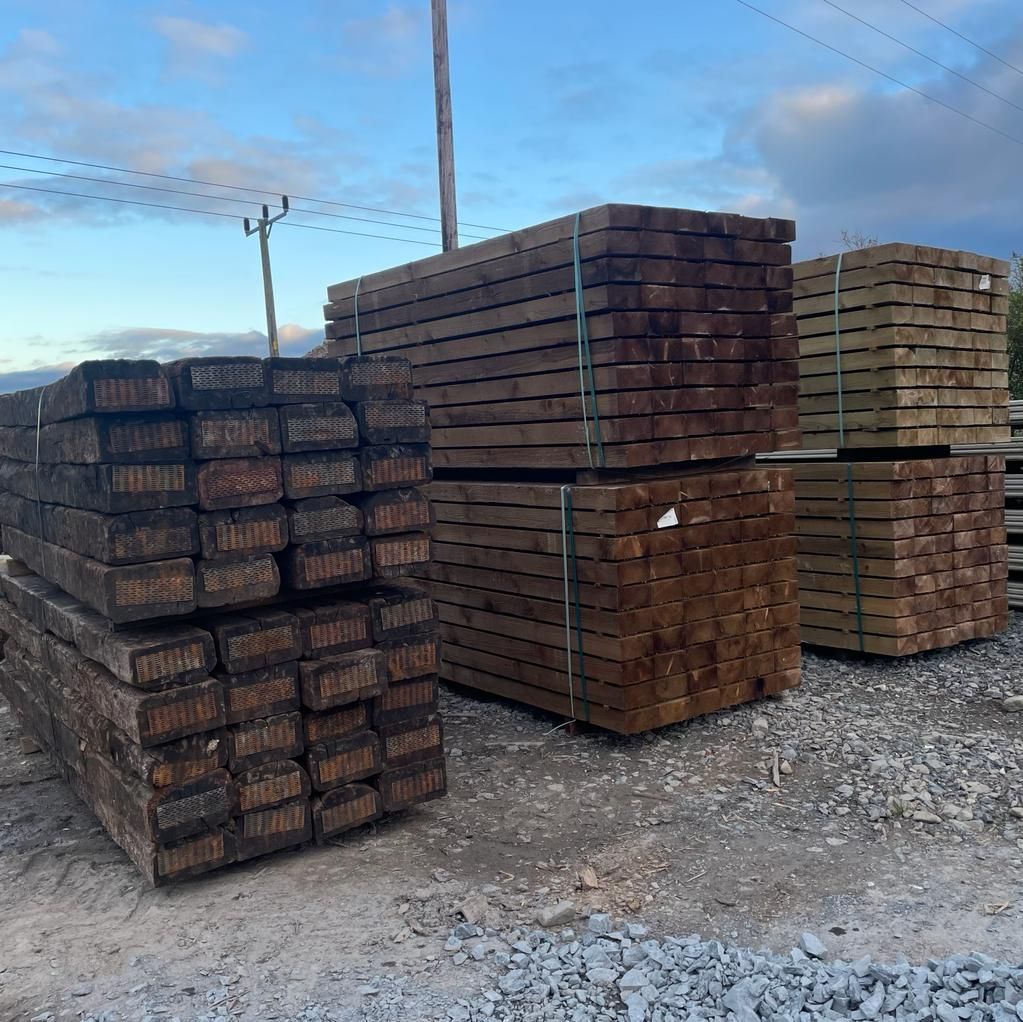 Large stacks of dark brown wooden railway sleepers bundled together in an outdoor yard with a gravel ground.