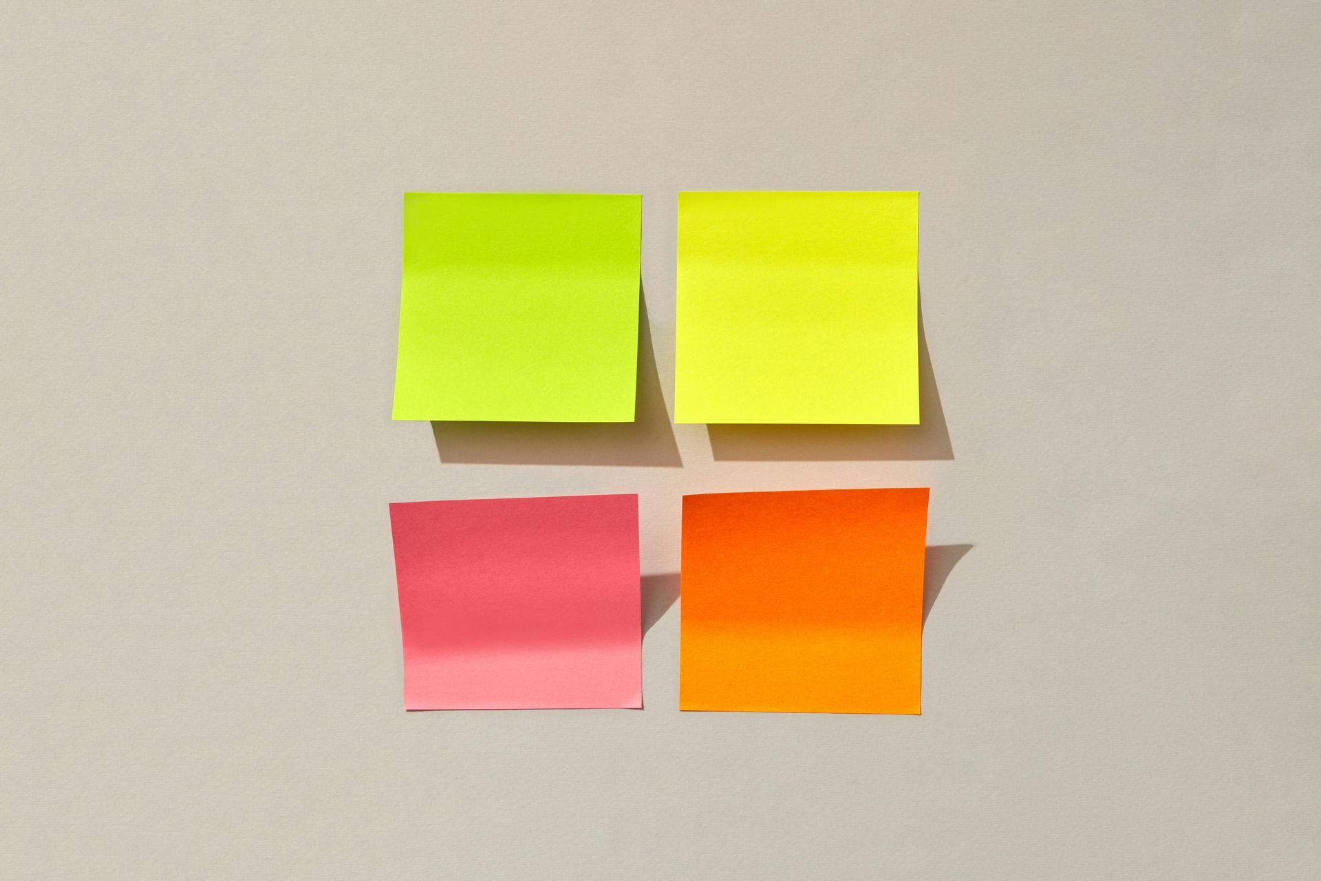 Four blank, square sticky notes in lime, yellow, pink, and orange arranged in a two-by-two grid against a beige background.