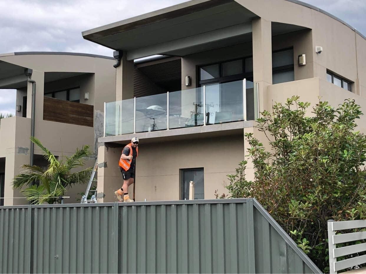 Worker standing on a ladder beside a modern house with a glass balcony and green fence