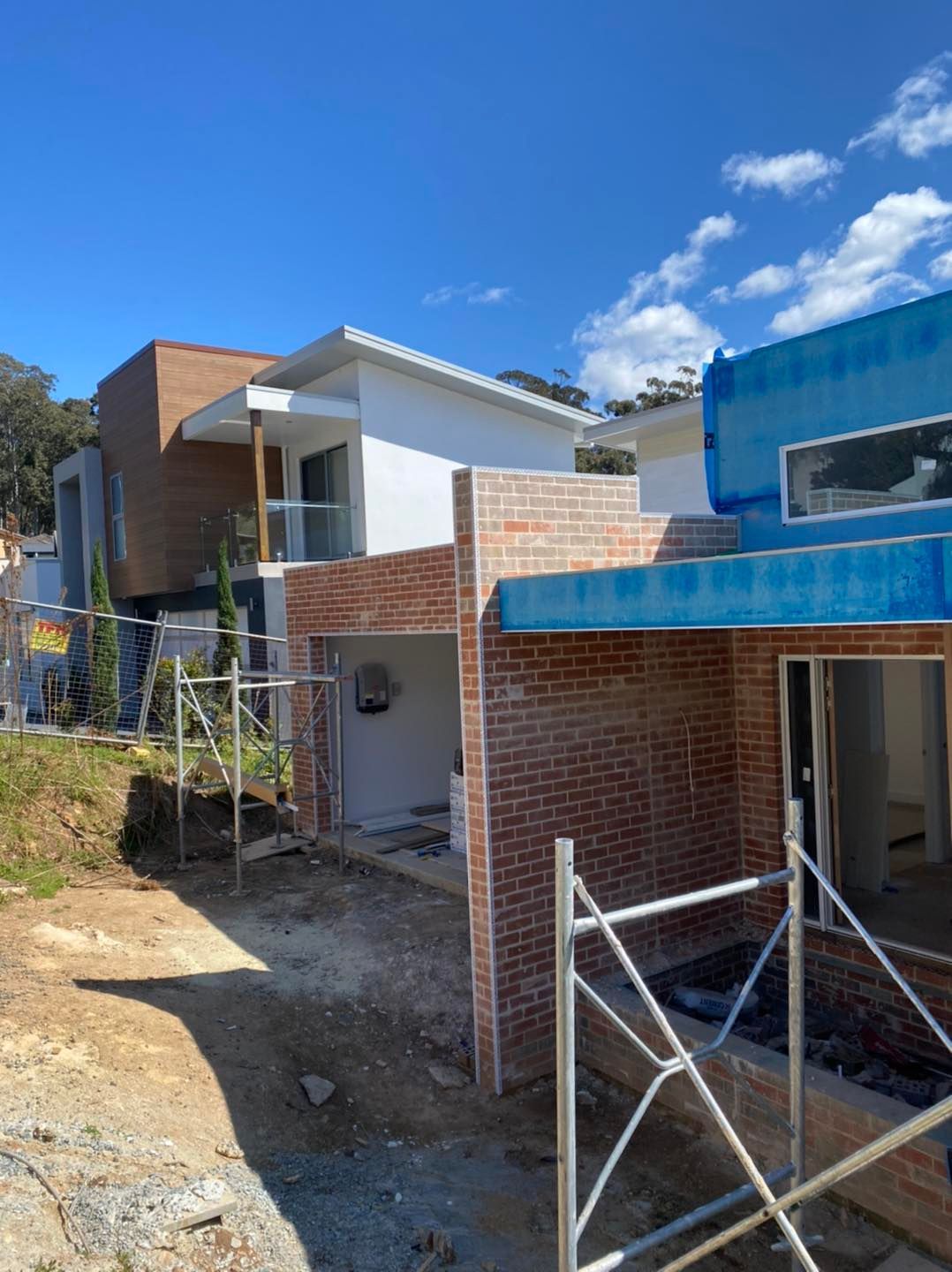 Partially built houses on a dirt lot under a blue sky, with brick, white, and blue exterior walls and scaffolding