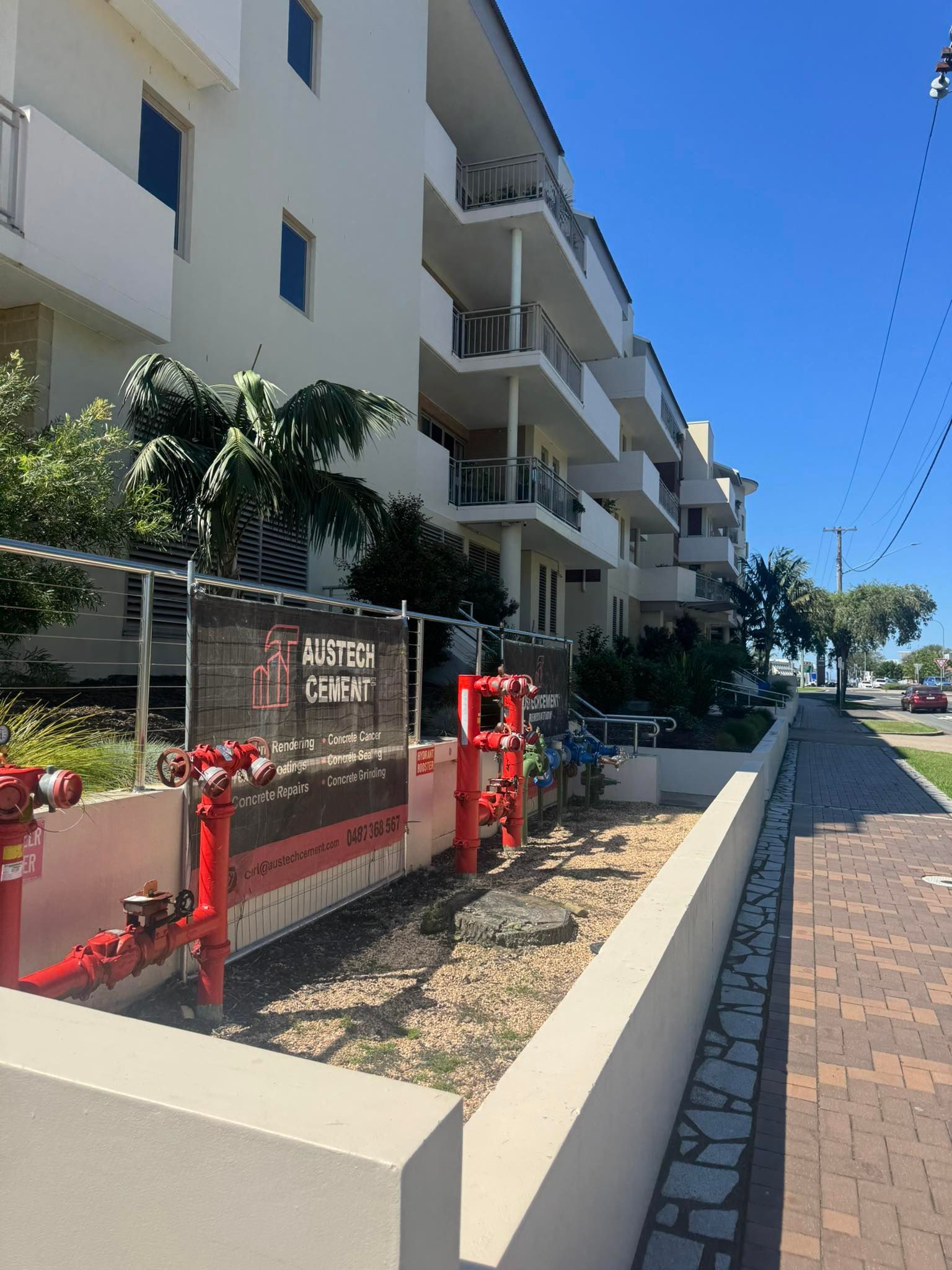 Apartment building beside a sunny sidewalk, with red utility equipment and a stone-lined path
