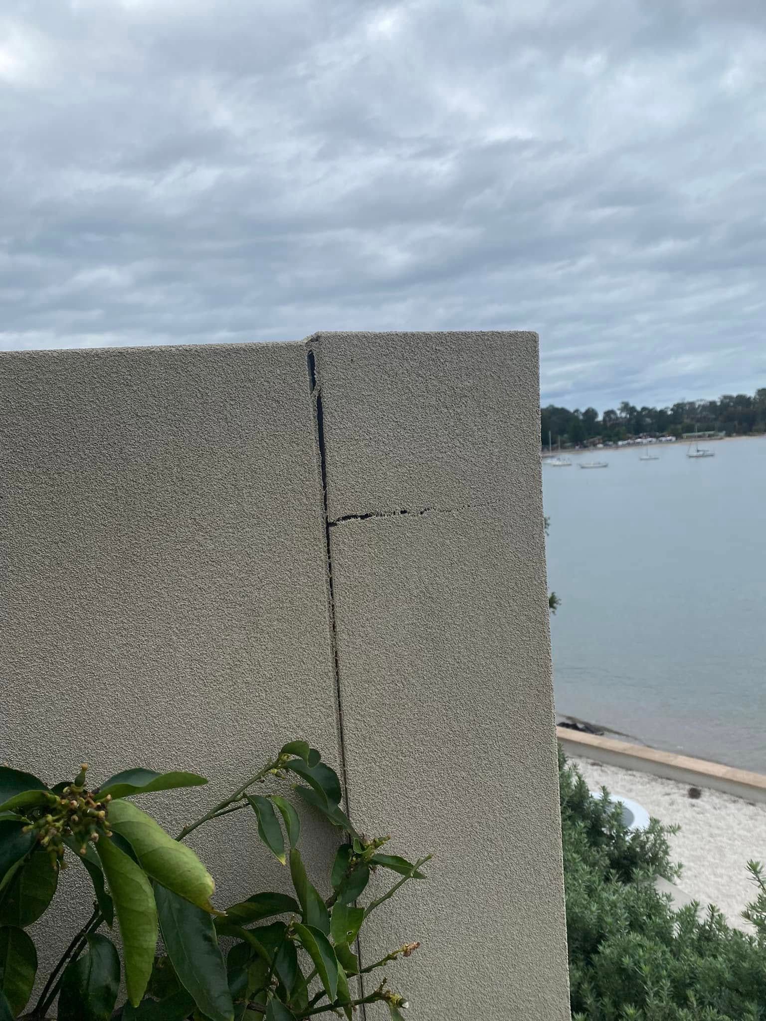 Cracked concrete wall beside a calm waterfront under cloudy skies, with green leaves in the foreground.