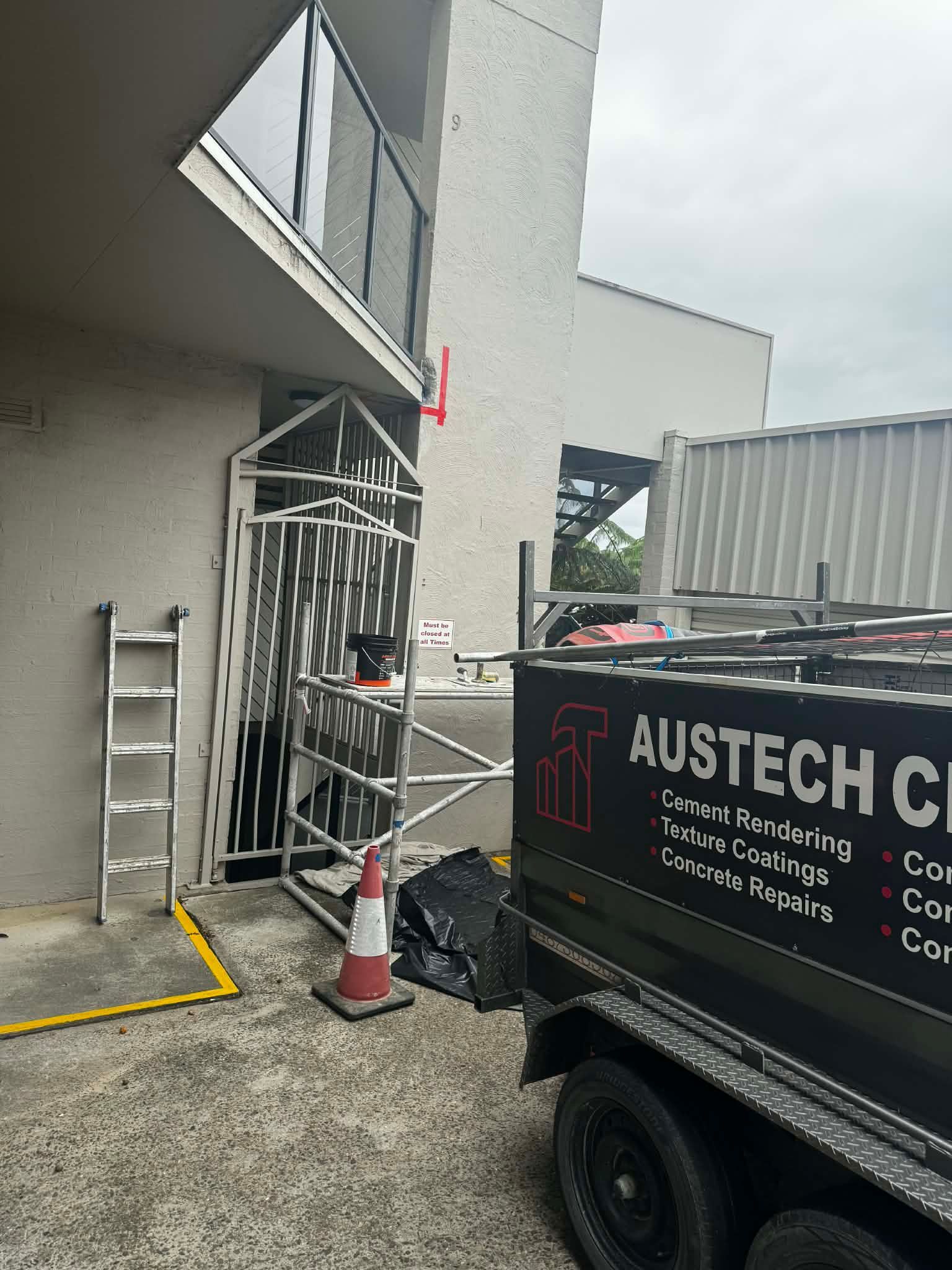 Construction area with white metal gate, cone, ladder, and black trailer beside a building.