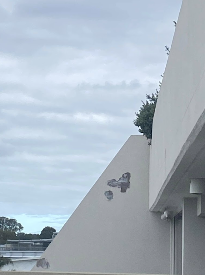 White building corner with a triangular wall, flaking render, mounted light, and cloudy sky over water in the distance