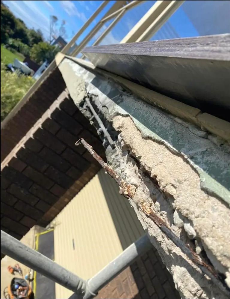 Close-up of a damaged building edge with crumbling concrete and exposed material under a roofline.