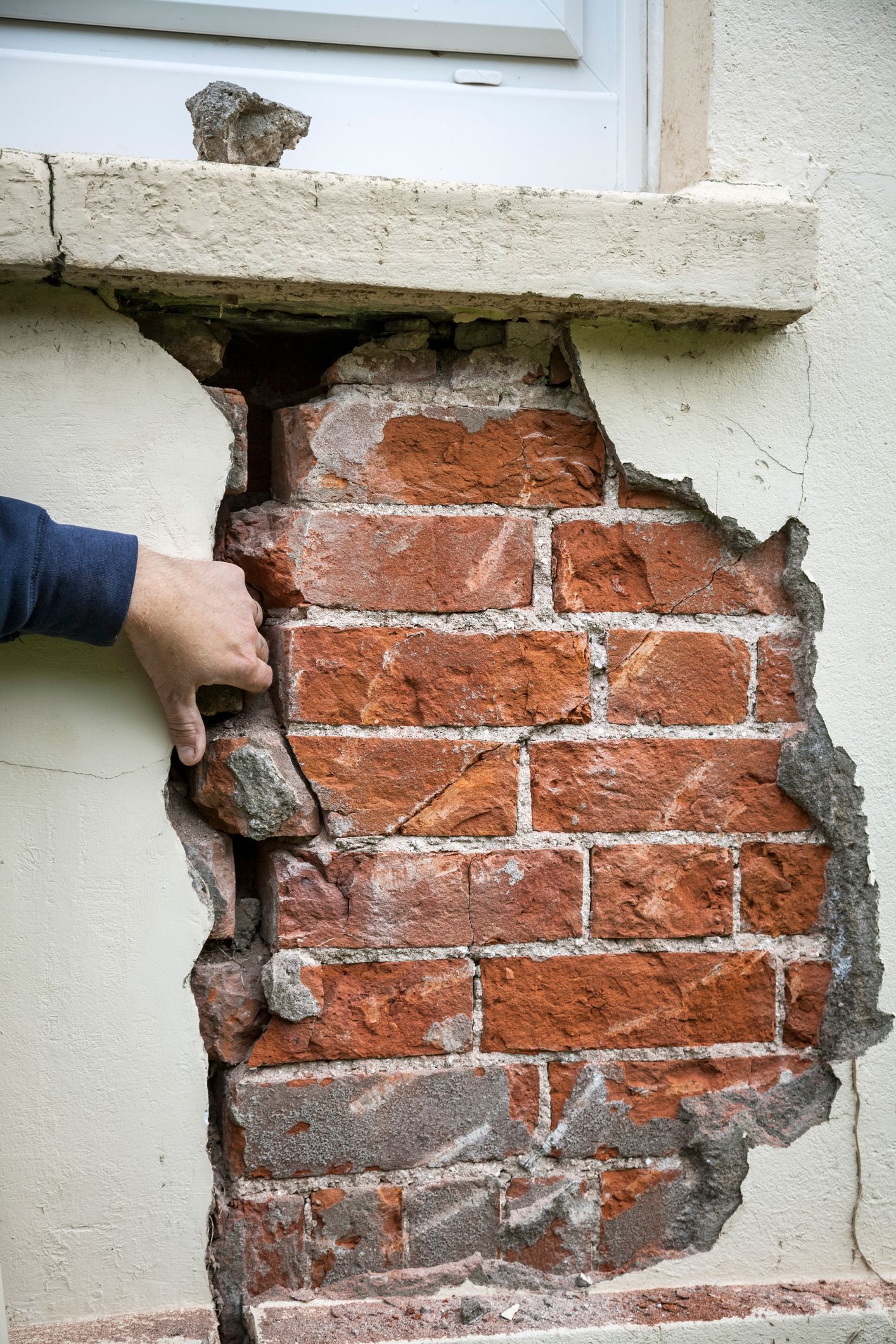 Hand exposing damaged red brick wall beneath cracked exterior plaster near a window