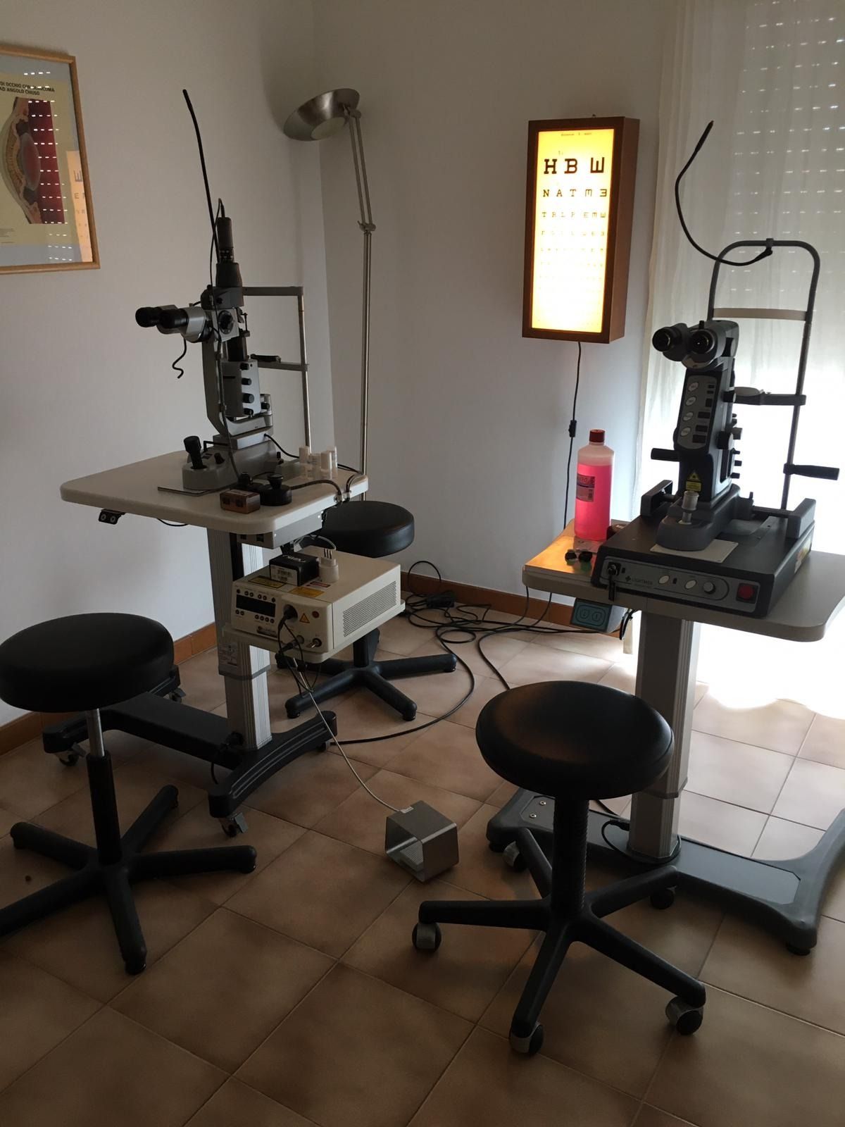 Due apparecchi per l'esame della vista in un ufficio con una tabella oculistica illuminata e sgabelli.