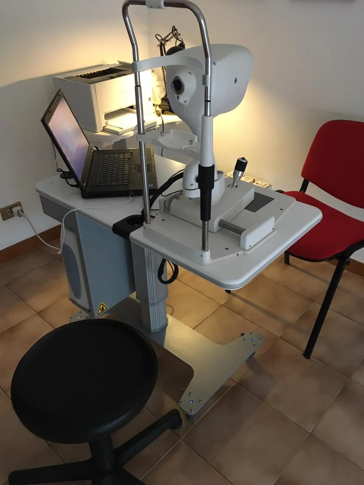 Attrezzatura per esami oculistici con computer portatile, stampante, sgabello e sedia nello studio di un medico.
