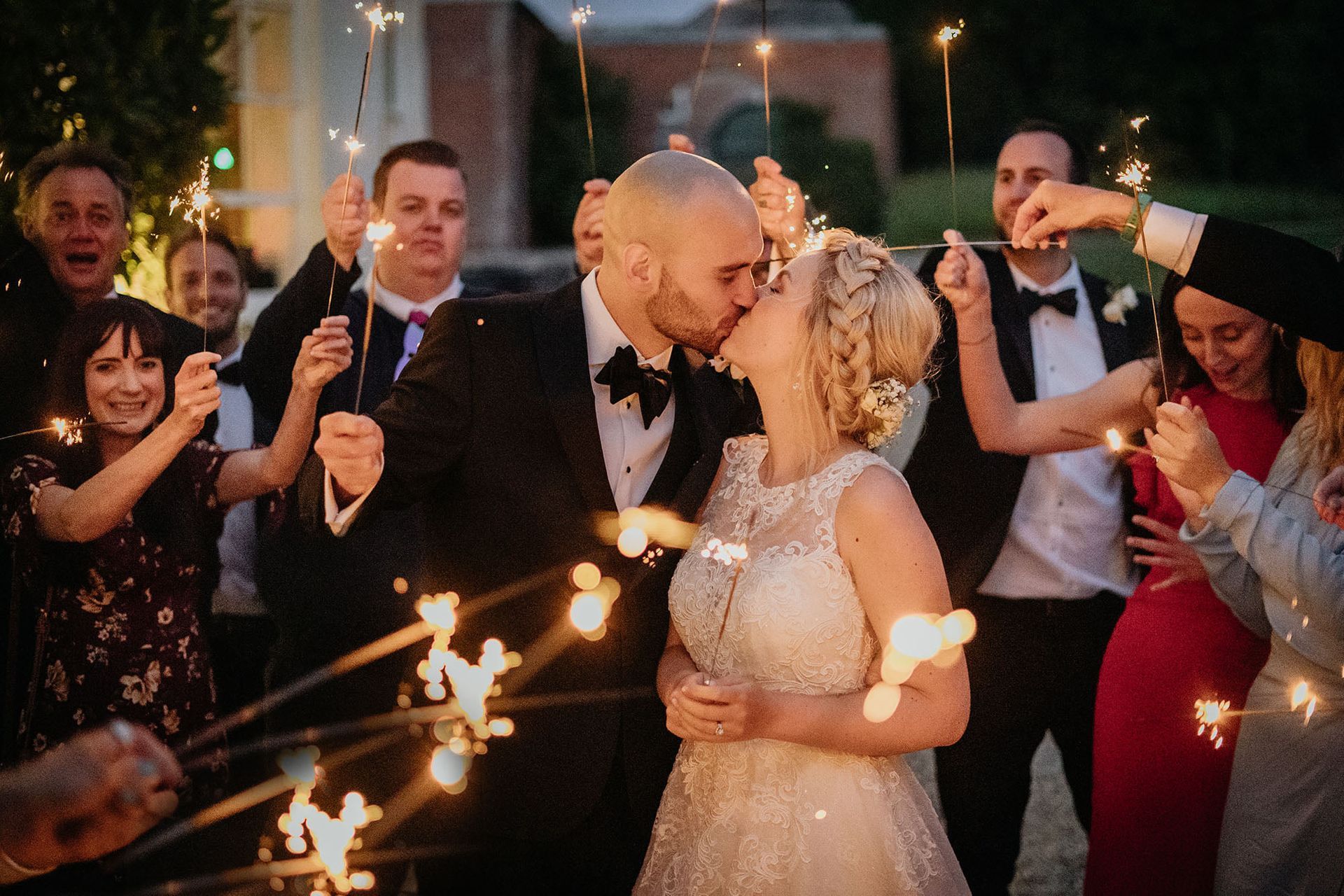 a bride and groom kissing while their wedding guests hold sparklers .