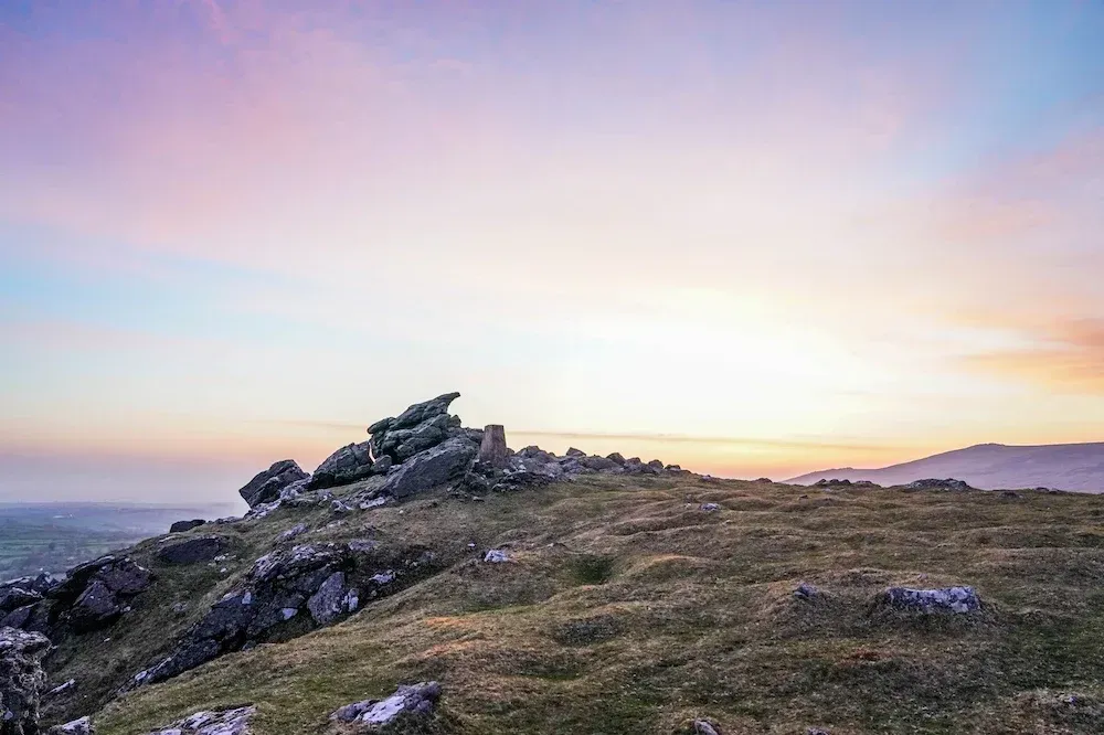 a mountain covered in grass and rocks with a sunset in the background.