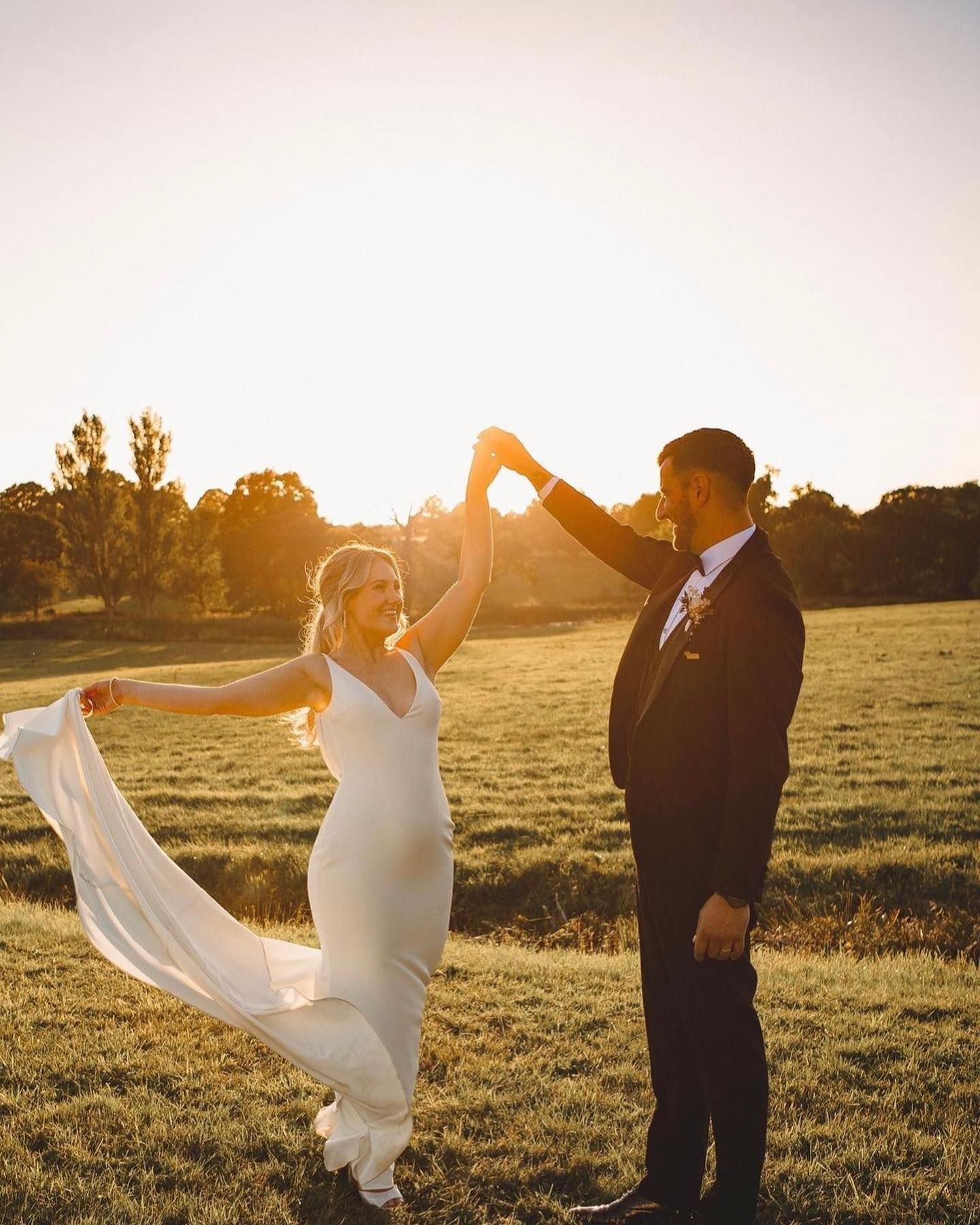 a bride and groom are dancing in a field at sunset .