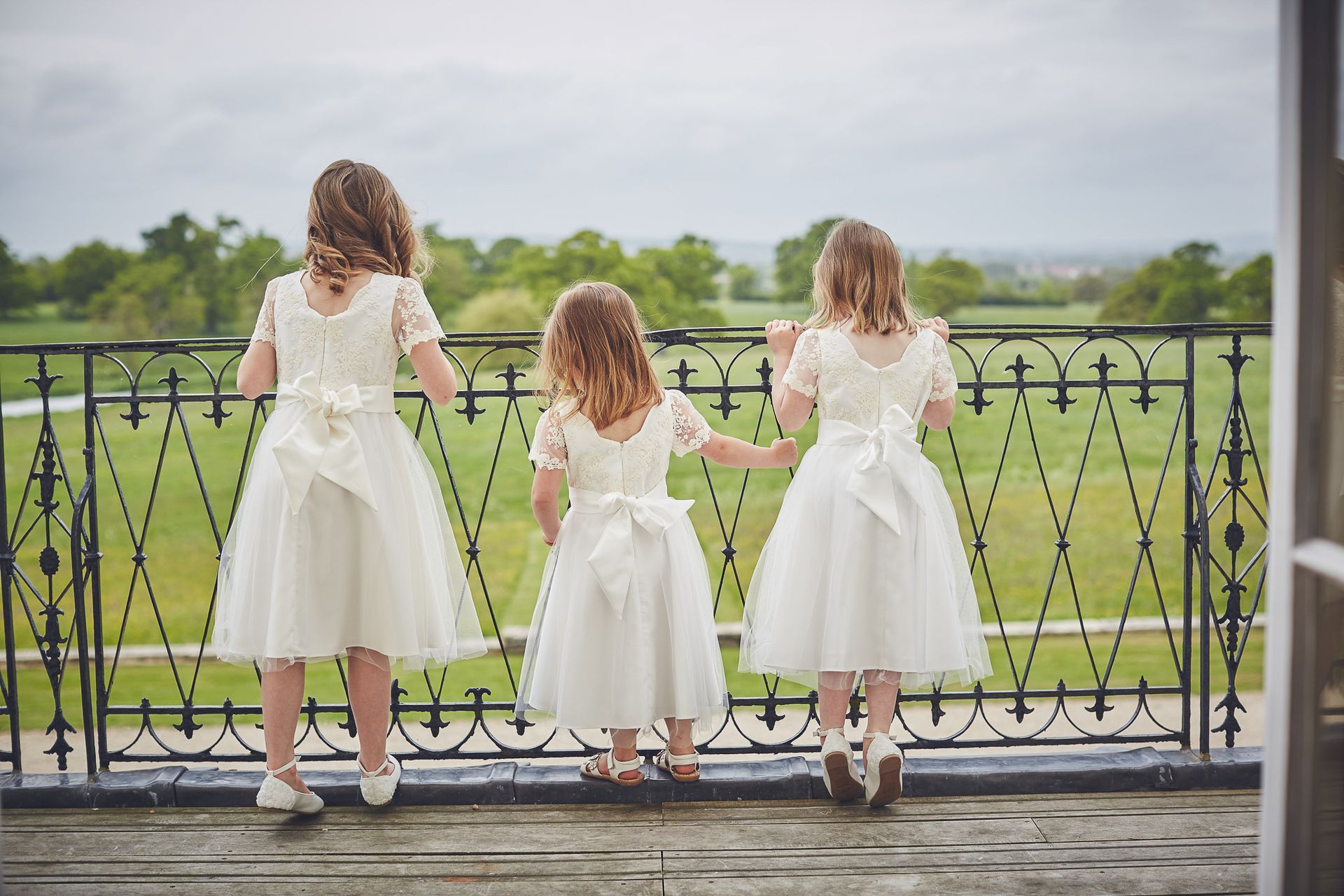 three flower girls in white dresses are standing on a balcony looking out over a field .