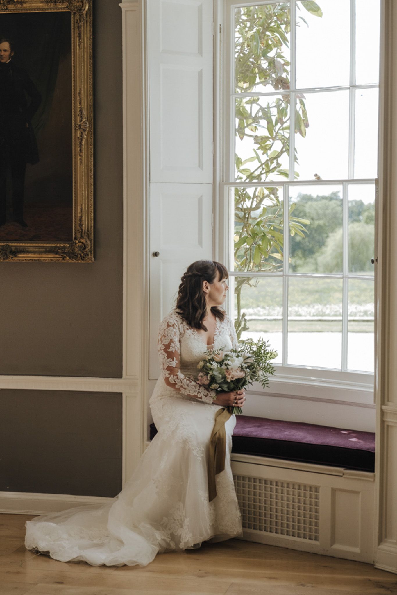 a bride in a wedding dress is sitting on a window sill holding a bouquet of flowers .
