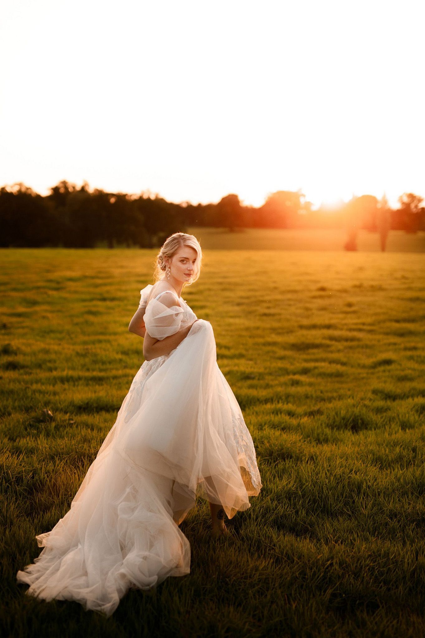 a woman in a wedding dress is standing in a field at sunset.