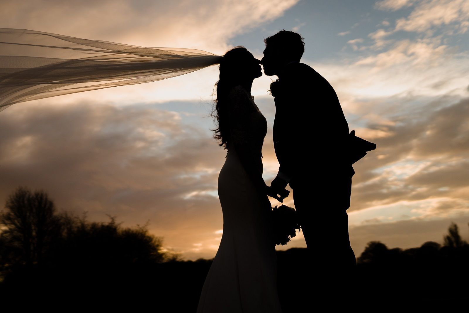a bride and groom kissing at sunset with the bride 's veil blowing in the wind .