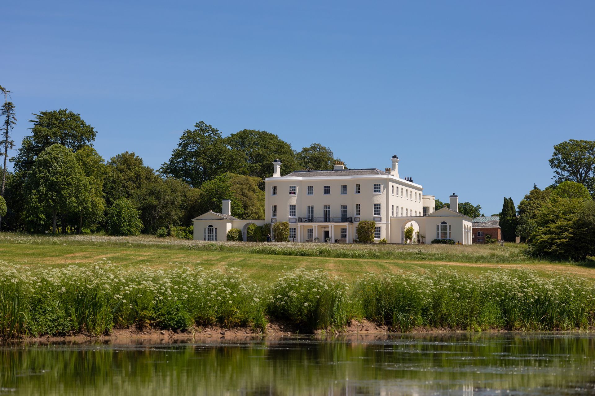 a large white house sits in the middle of a grassy field next to a body of water. Wedding venue in Devon.