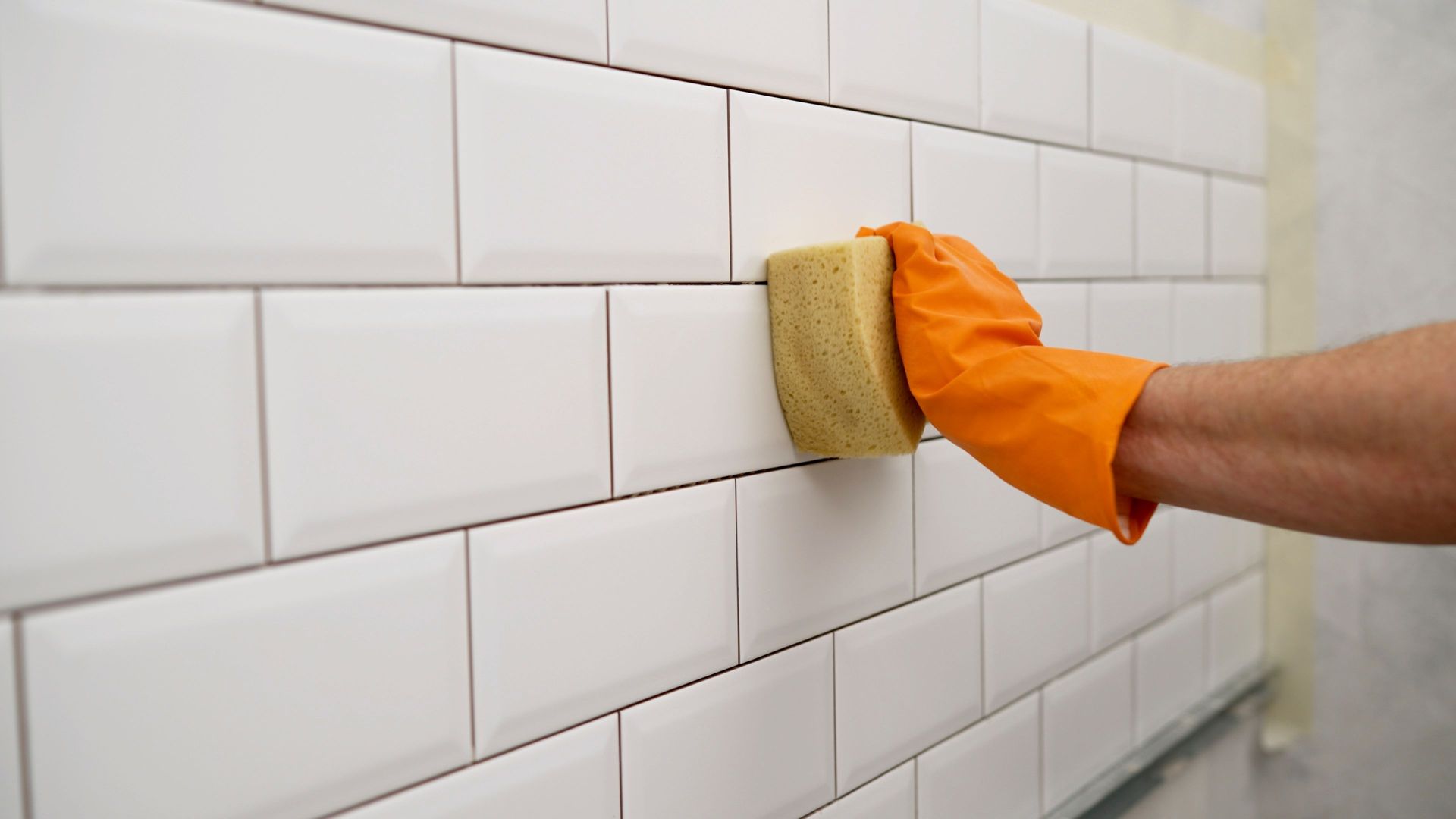 Gloved hand scrubbing white tiled wall with a sponge