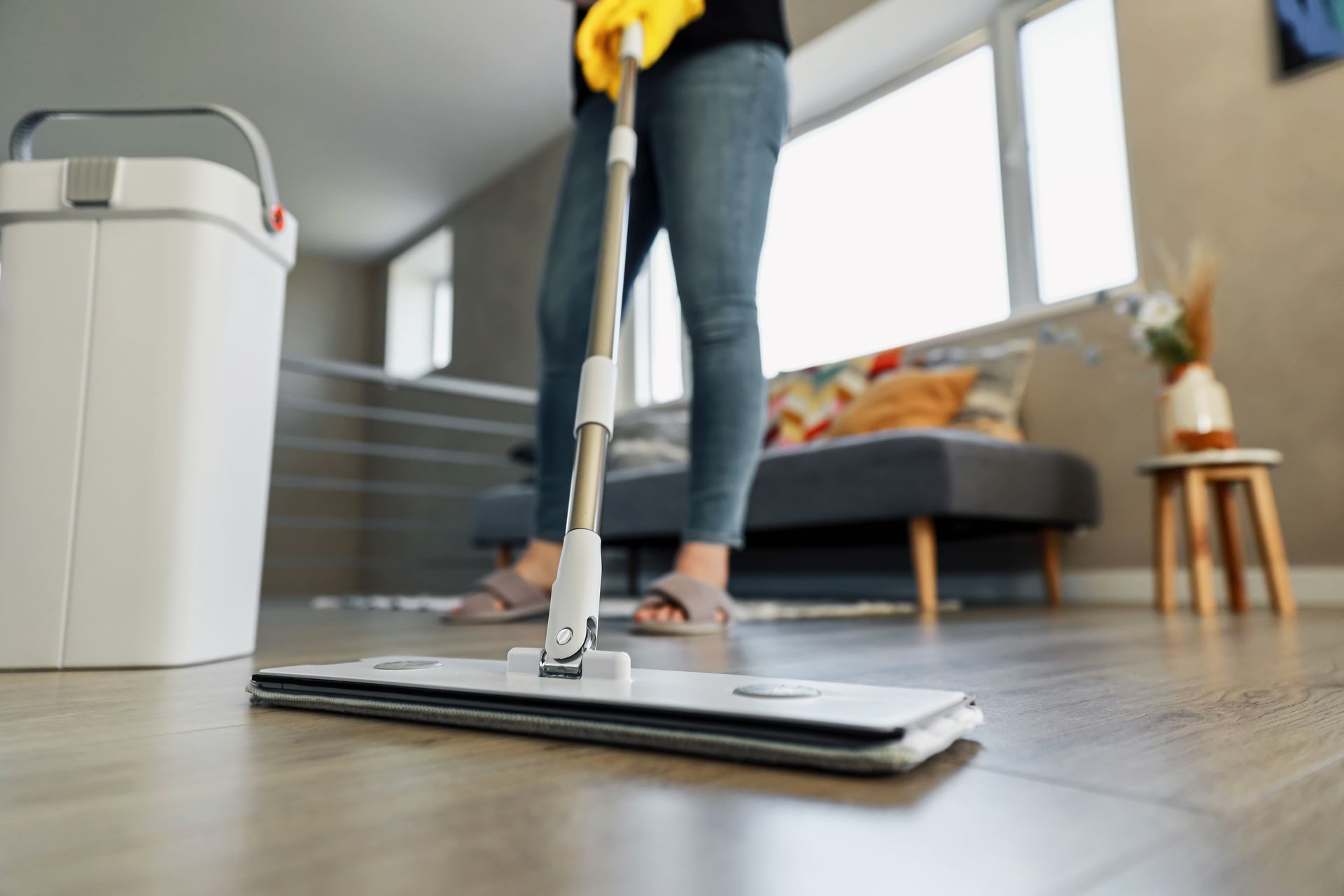 Person mopping a wooden floor in a bright living room with a white bucket nearby