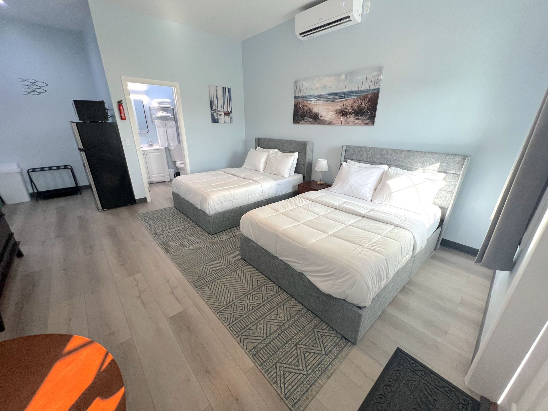 Bedroom with two beds, light blue walls, art, a rug, and a bathroom doorway.