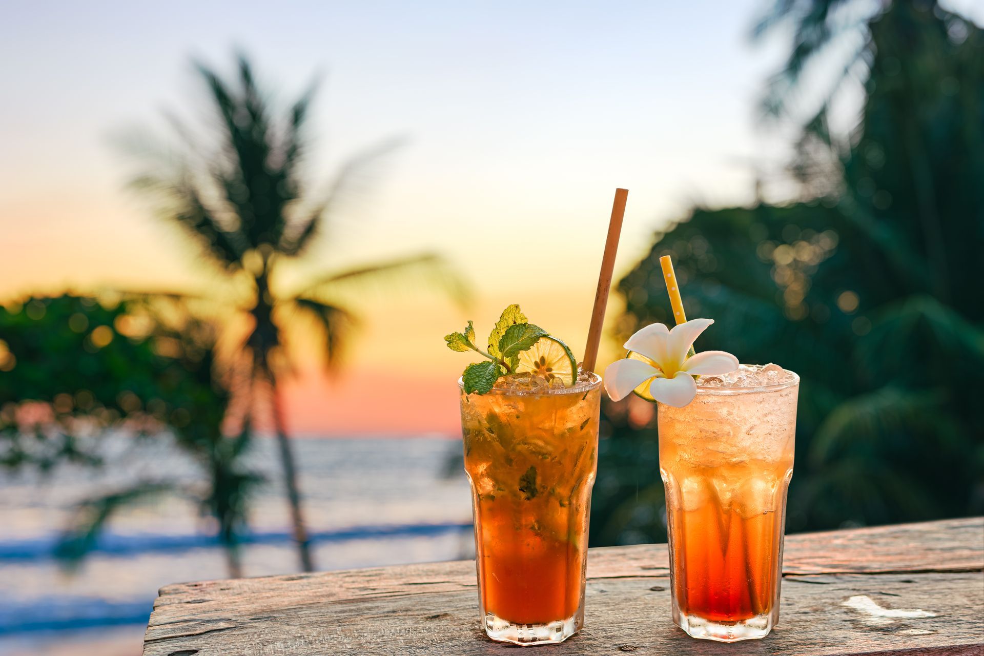 Two iced cocktails with straws and garnishes on a wooden surface, beach and sunset in the background.