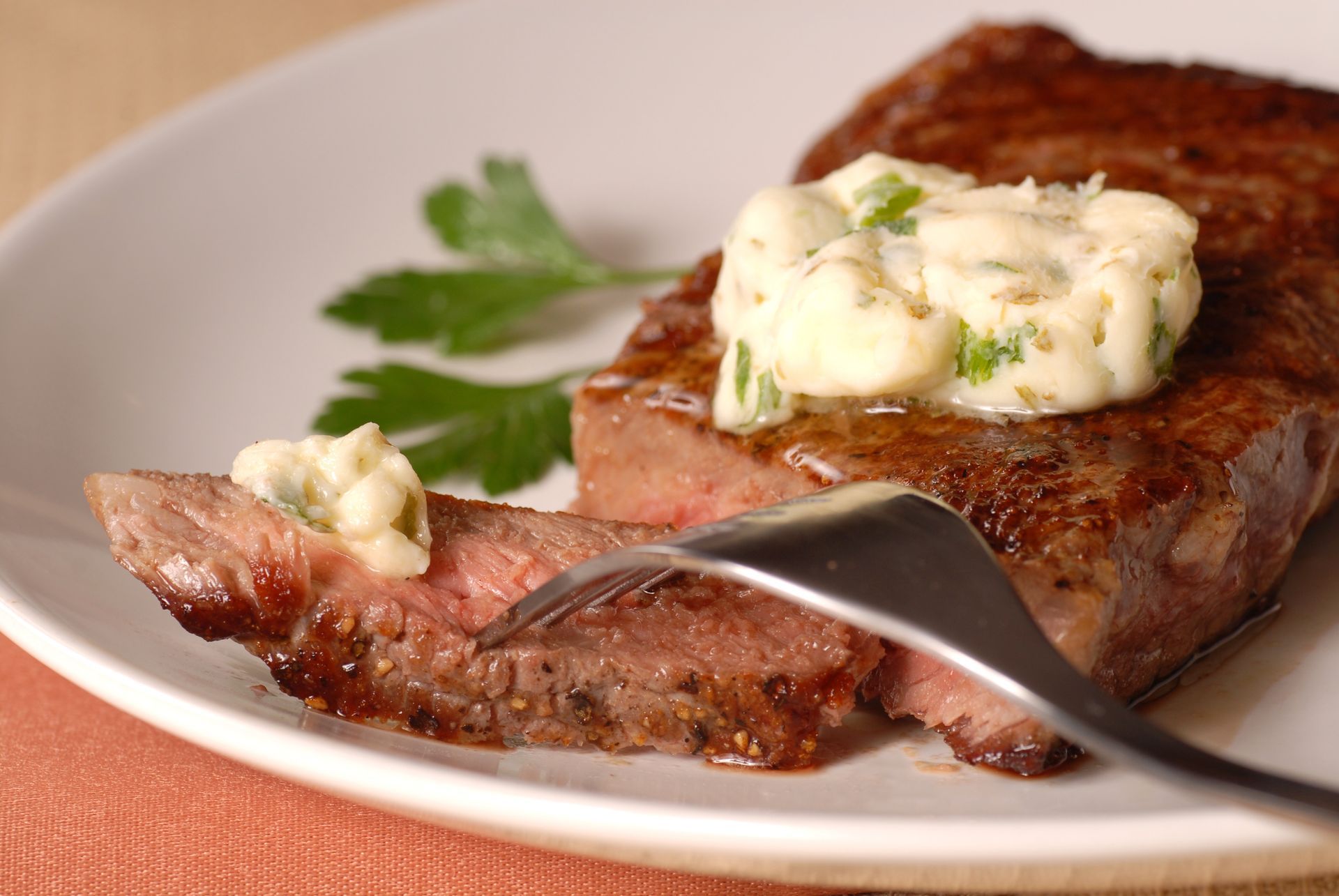 Grilled steak with herb butter on white plate; a fork slices a piece, showing pink inside.