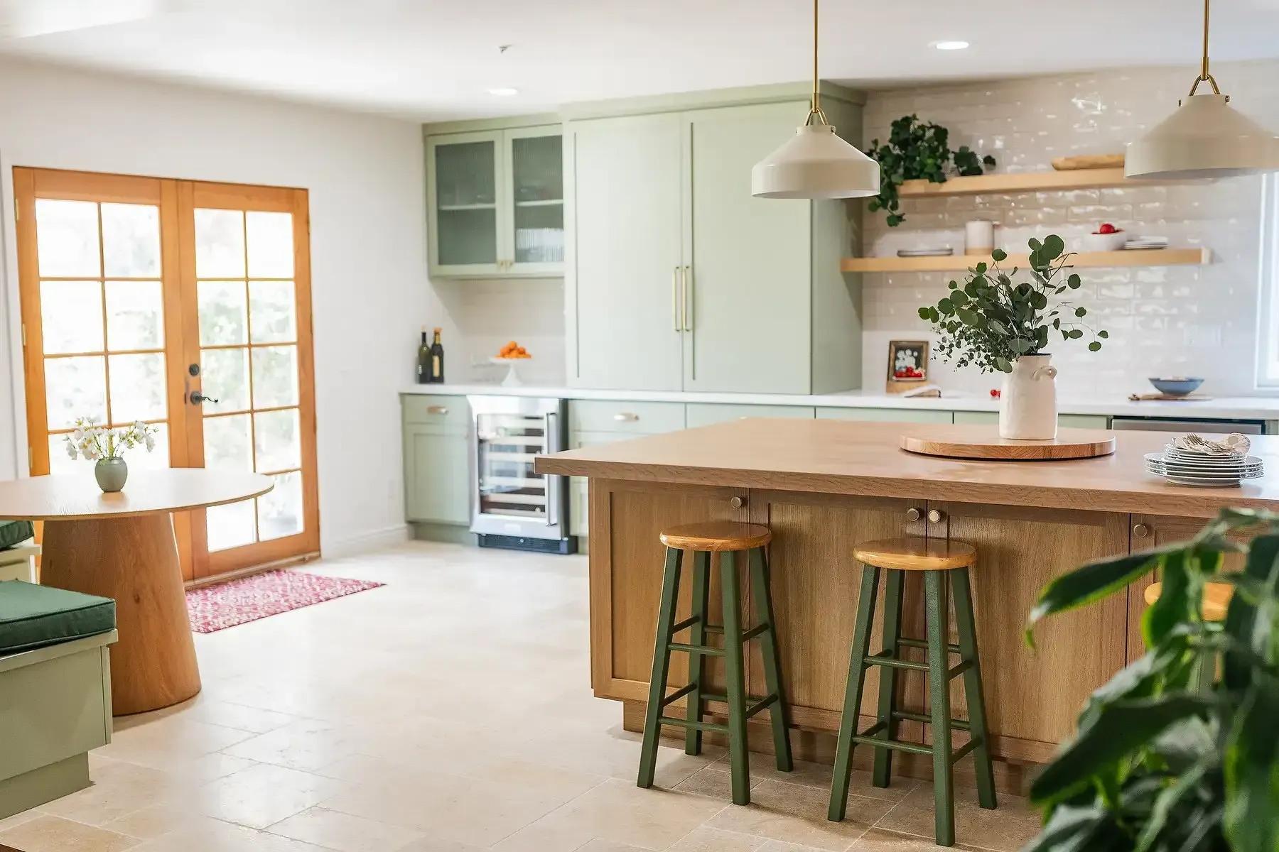 Kitchen with sage green cabinets, light wood island and table, stools, plants, and French doors.