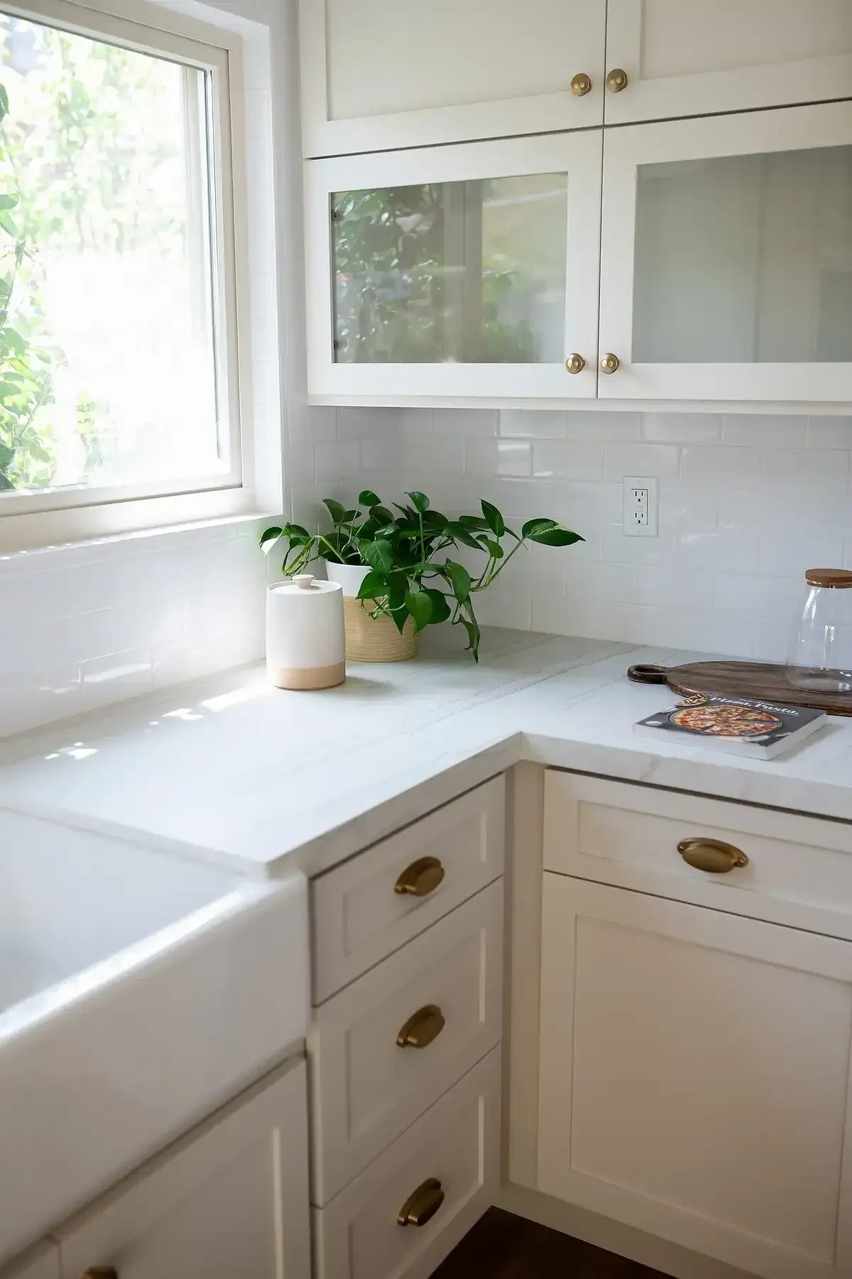 Team Renovate - White kitchen with farmhouse sink, cabinets, marble countertop, brass hardware, and window with greenery.