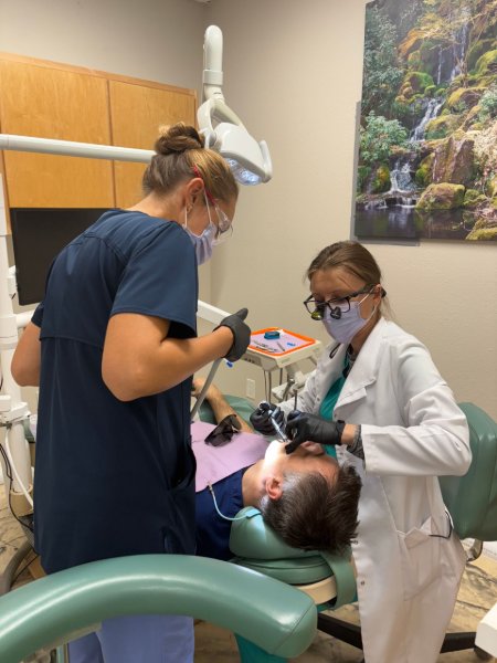 Dentist and assistant treating a patient in a dental exam room, with equipment and a monitor nearby