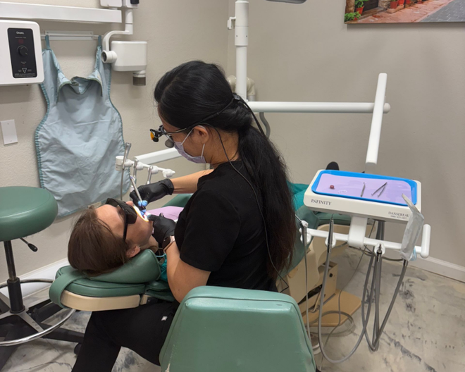 Dental hygienist working on a patient in a dental chair, with a monitor and equipment nearby.