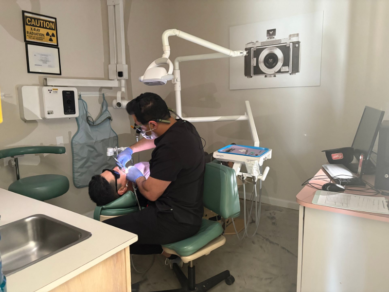 Dentist treating a patient in a dental clinic, with exam light and equipment visible.
