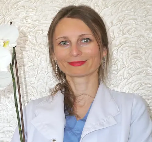 Woman in white coat and blue top, smiling against a textured white background.