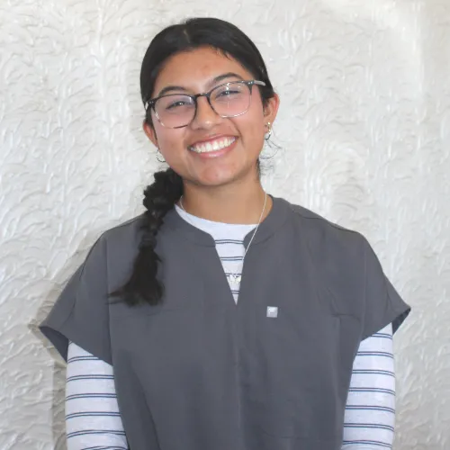 Smiling person with glasses and braided hair, wearing a gray top over a striped shirt against a white textured background