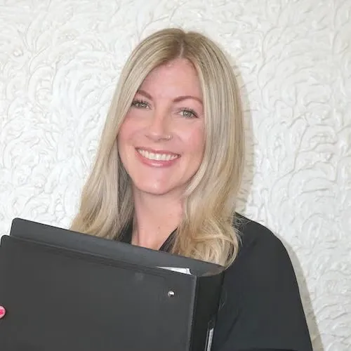 Smiling blonde woman holding a black binder against a white textured wall