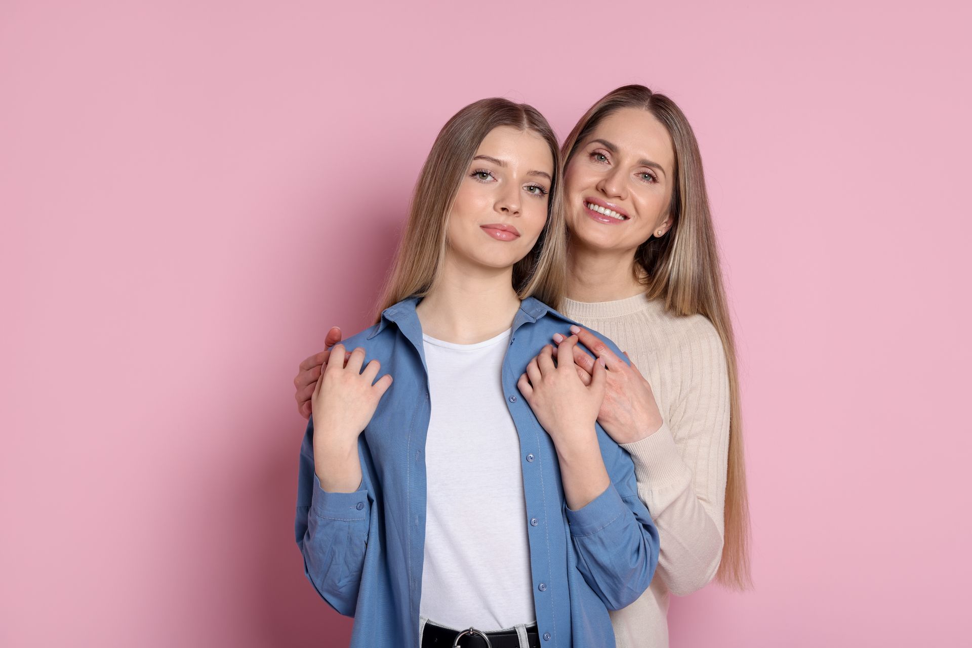 Two smiling women standing together against a pink background, one resting hands on the other's shoulders.
