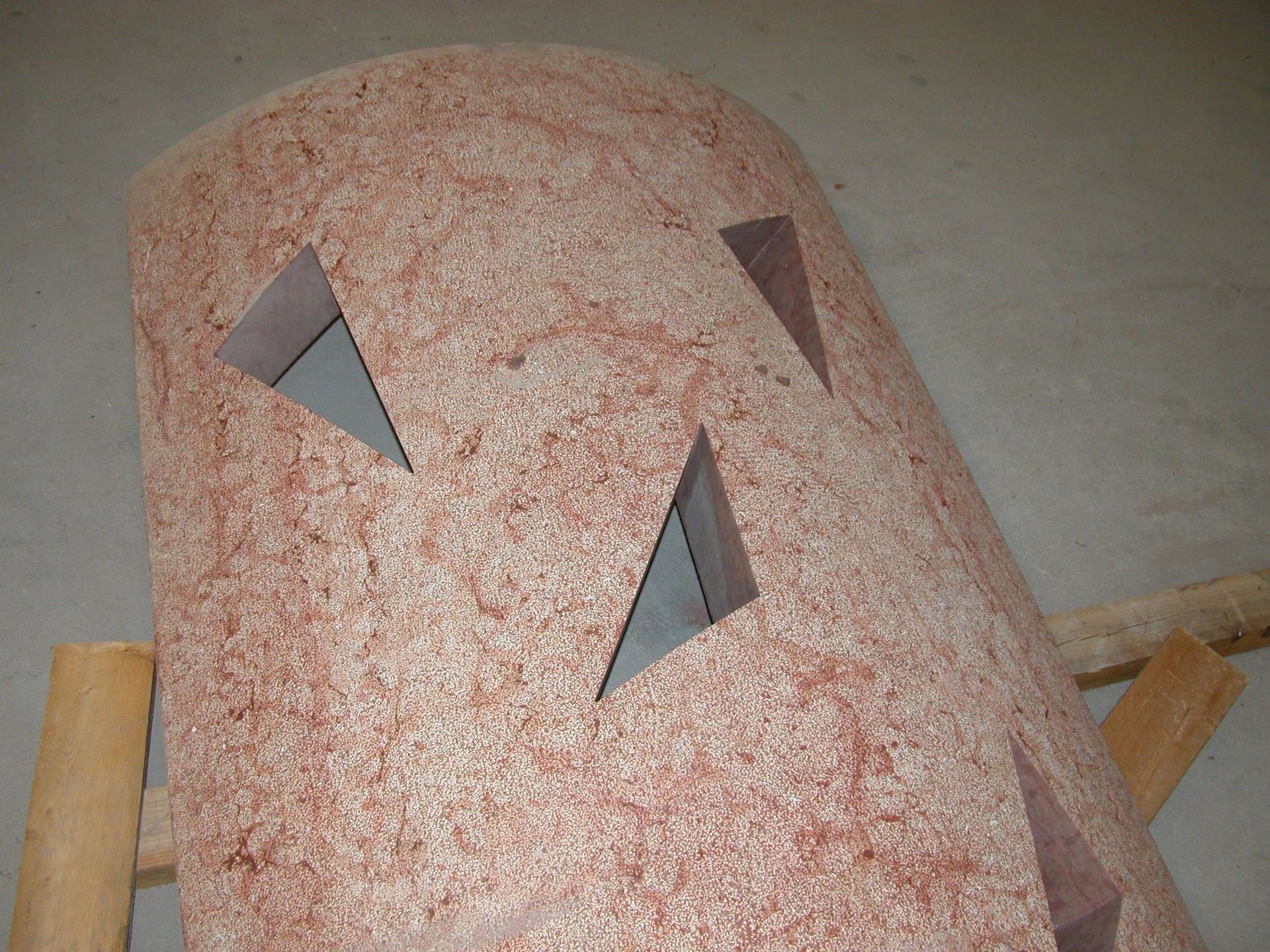 Oggetto a forma di cupola con macchie rosa e tre aperture triangolari, poggiante su un supporto di legno.
