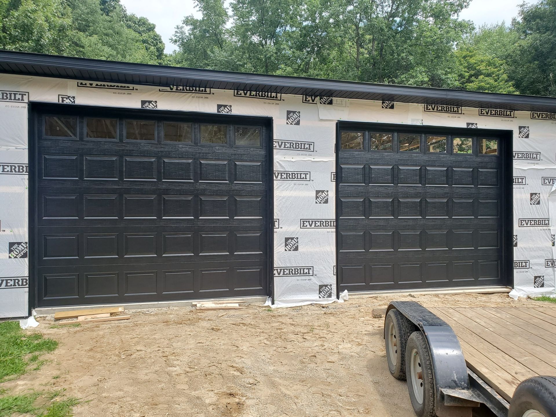 Two black garage doors on a house under construction with exposed house wrap and a trailer in the foreground