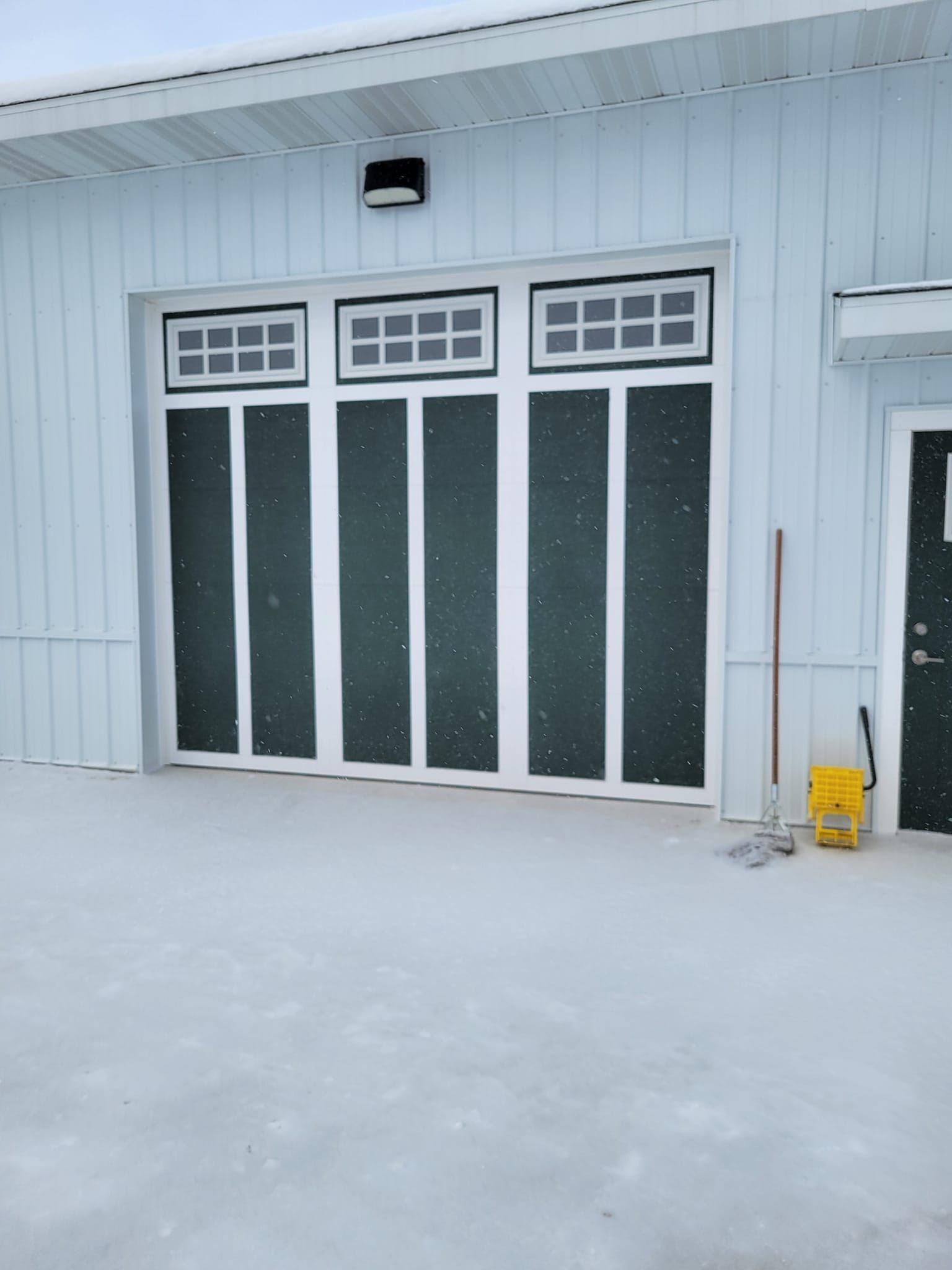 Snowy house exterior with white siding, a frosted front door, and a yellow snow shovel by the wall