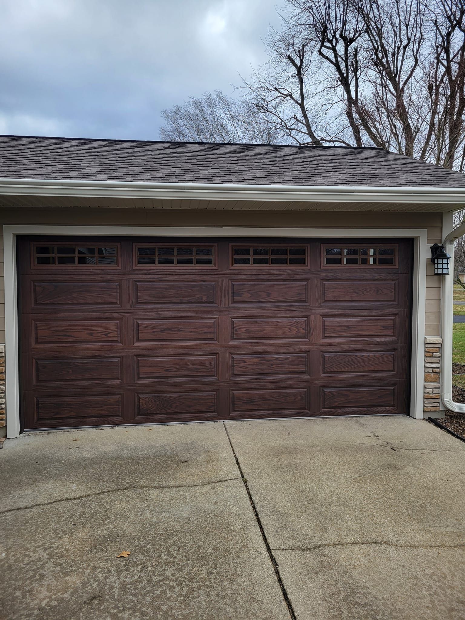 Brown double garage door under a gray roof with a concrete driveway and bare trees overhead