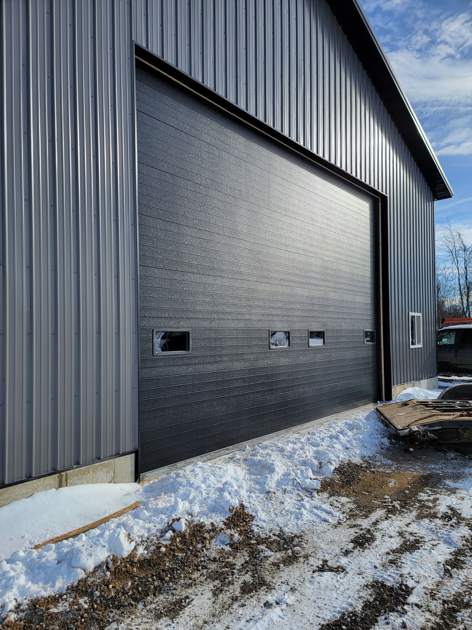 Black metal garage door on a corrugated gray industrial building with snow on the ground