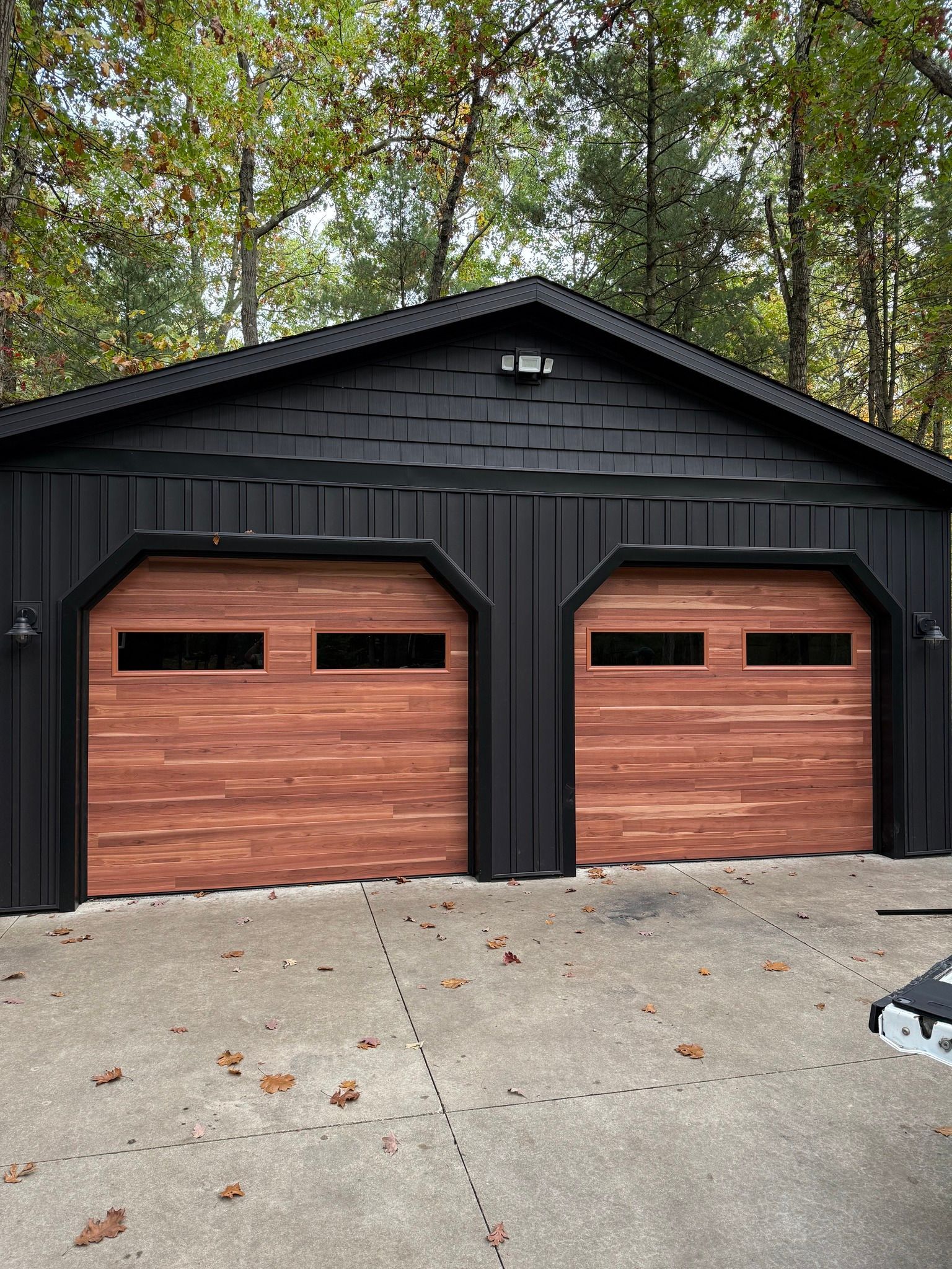 Black two-car garage with wood doors and a concrete driveway, surrounded by trees