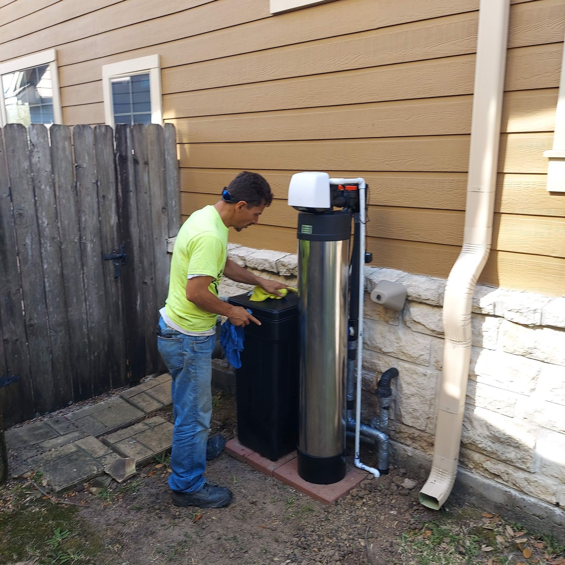 Man installing a water filtration system outside a house. Brown siding, silver tank, and white piping.